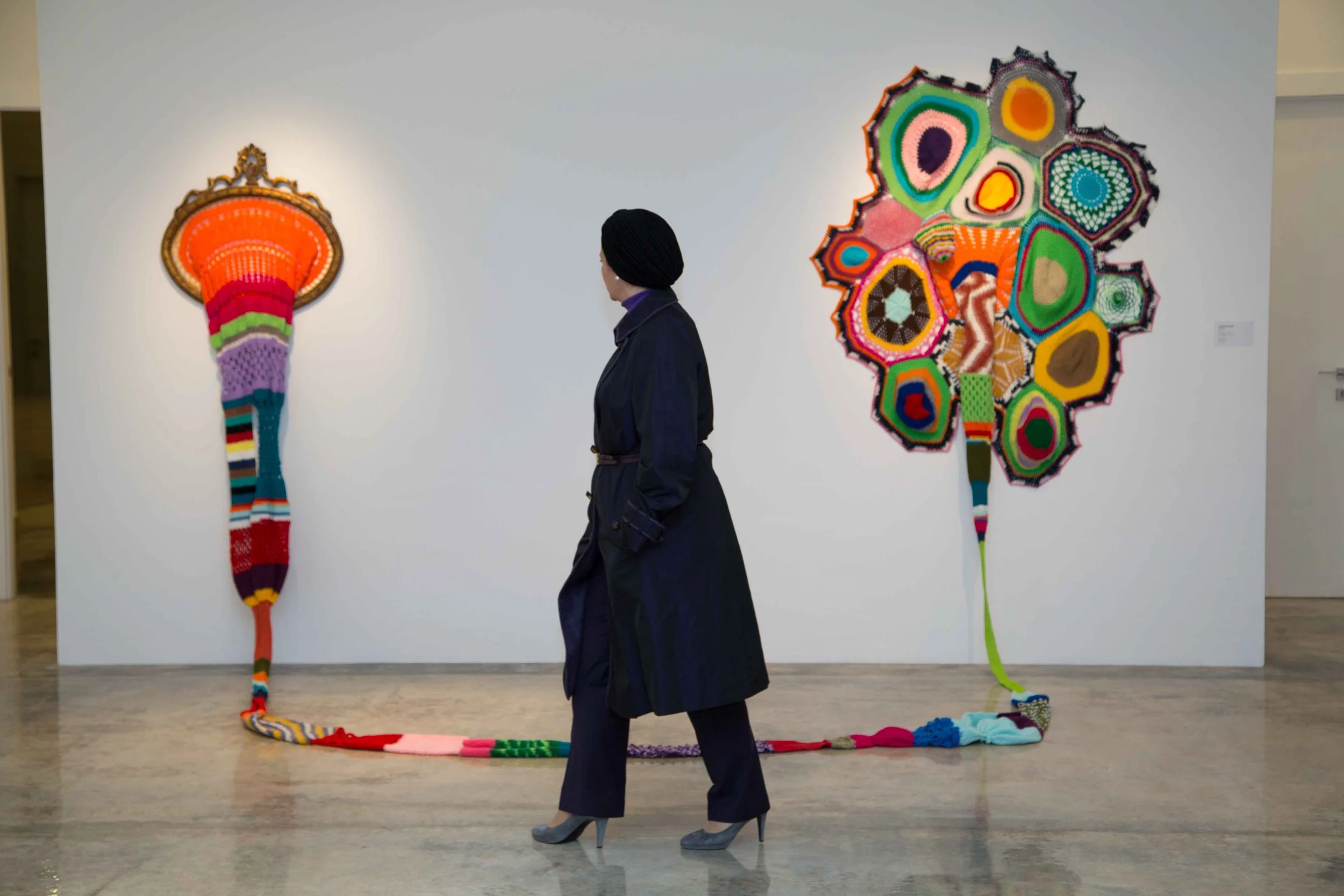 A woman in a black coat and heels standing in front of colorful knitted art pieces on a gallery wall, including a tall, rainbow-colored, elongated piece on the left and a multicolored abstract shape on the right.