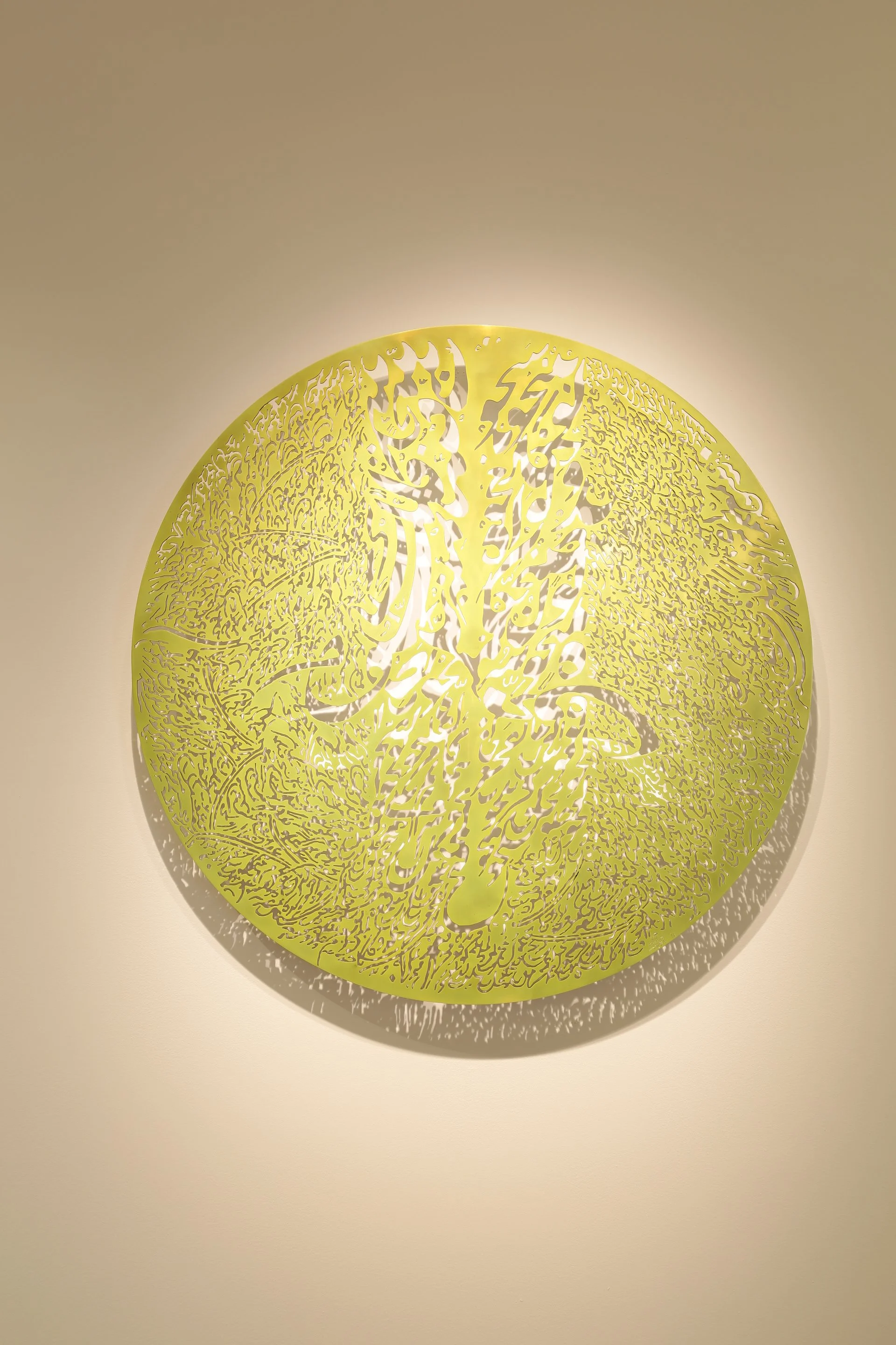 A round, yellow metal artwork with intricate Arabic calligraphy and designs, casting shadows on a light-colored wall.