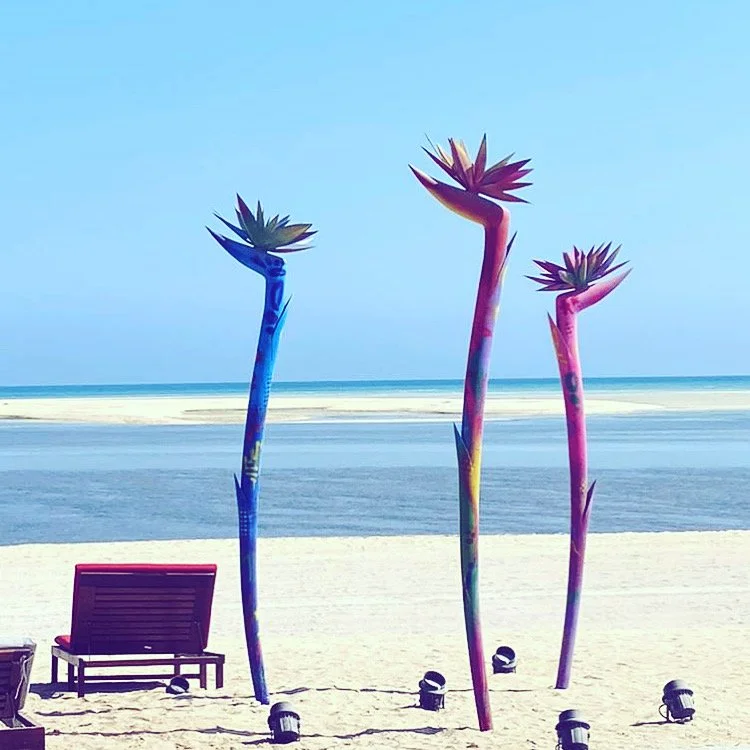 Colorful artificial plants resembling succulents on a sandy beach with lounge chairs and ocean in the background.