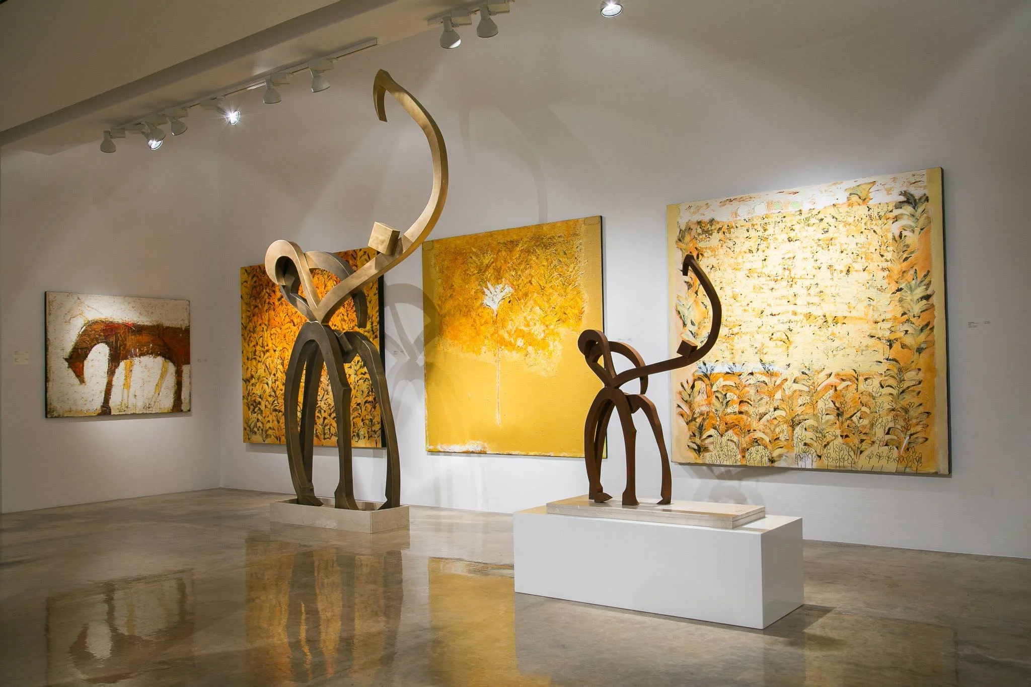 Art gallery with abstract paintings and sculptures, including two large metal sculptures resembling stylized animals, displayed against a white wall with track lighting.