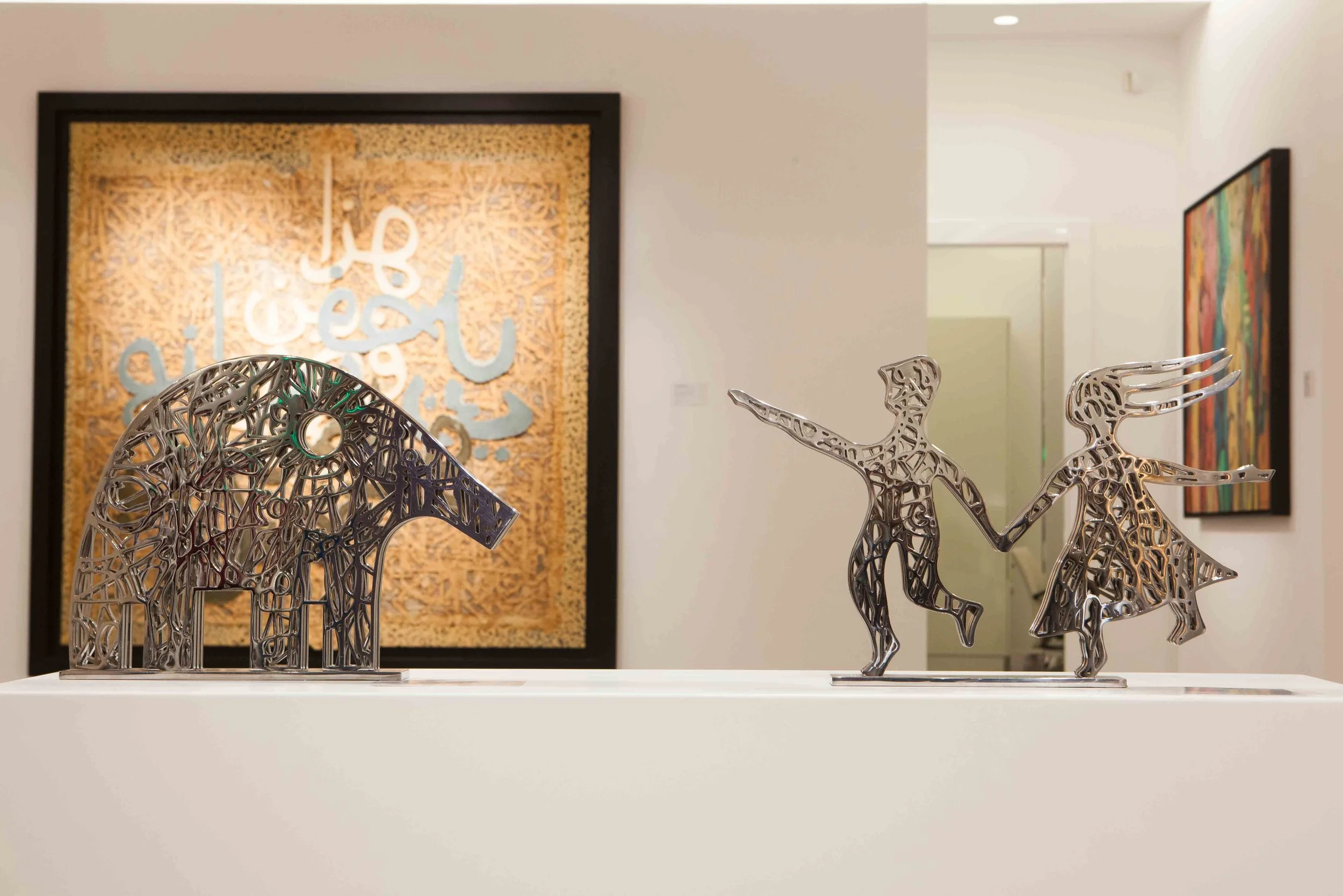 Metal art sculptures of an elephant and a dancing couple displayed on a white pedestal in an art gallery with framed abstract and textual artworks in the background.