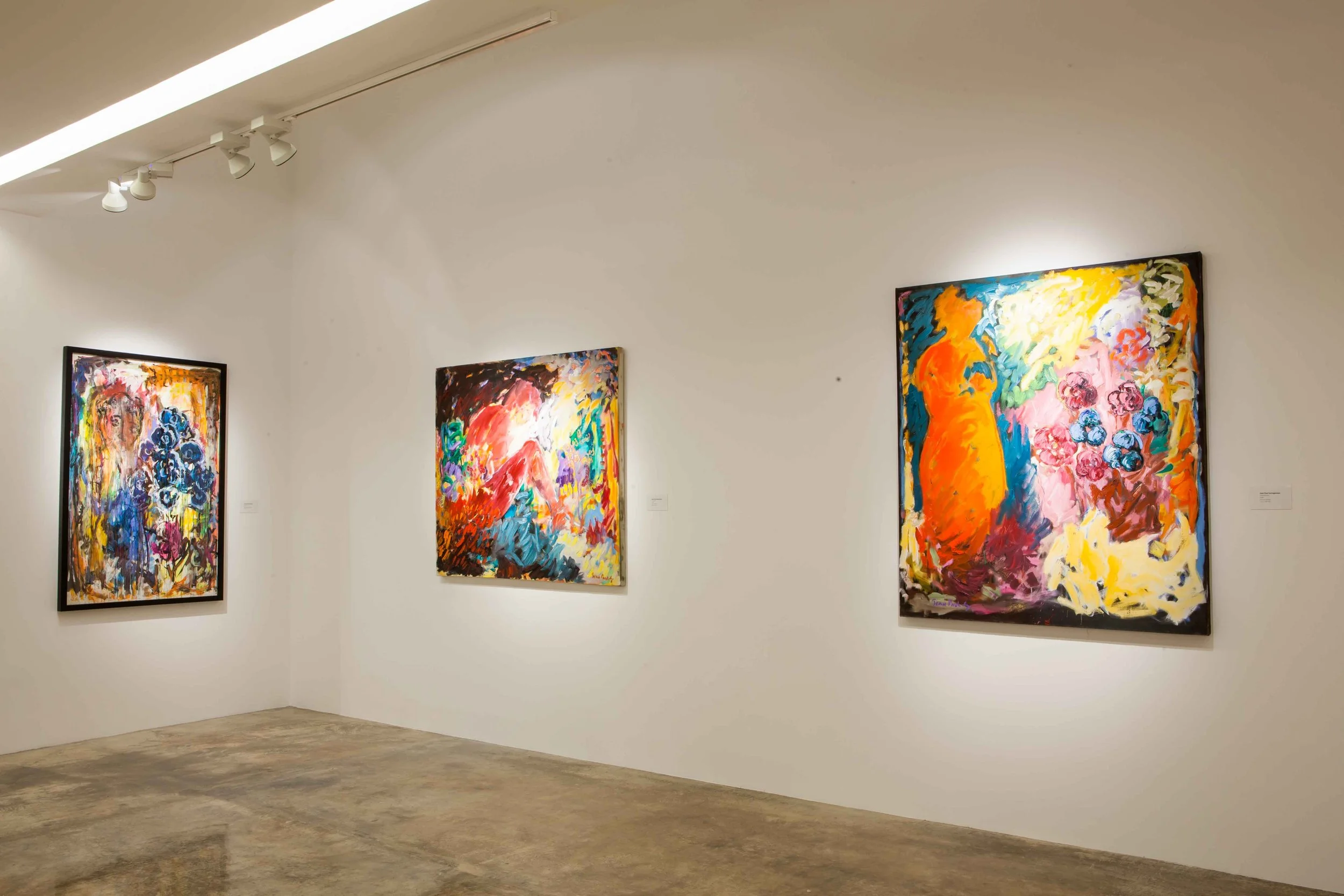 Three colorful abstract paintings on a gallery wall, illuminated by ceiling lights, with a polished concrete floor.
