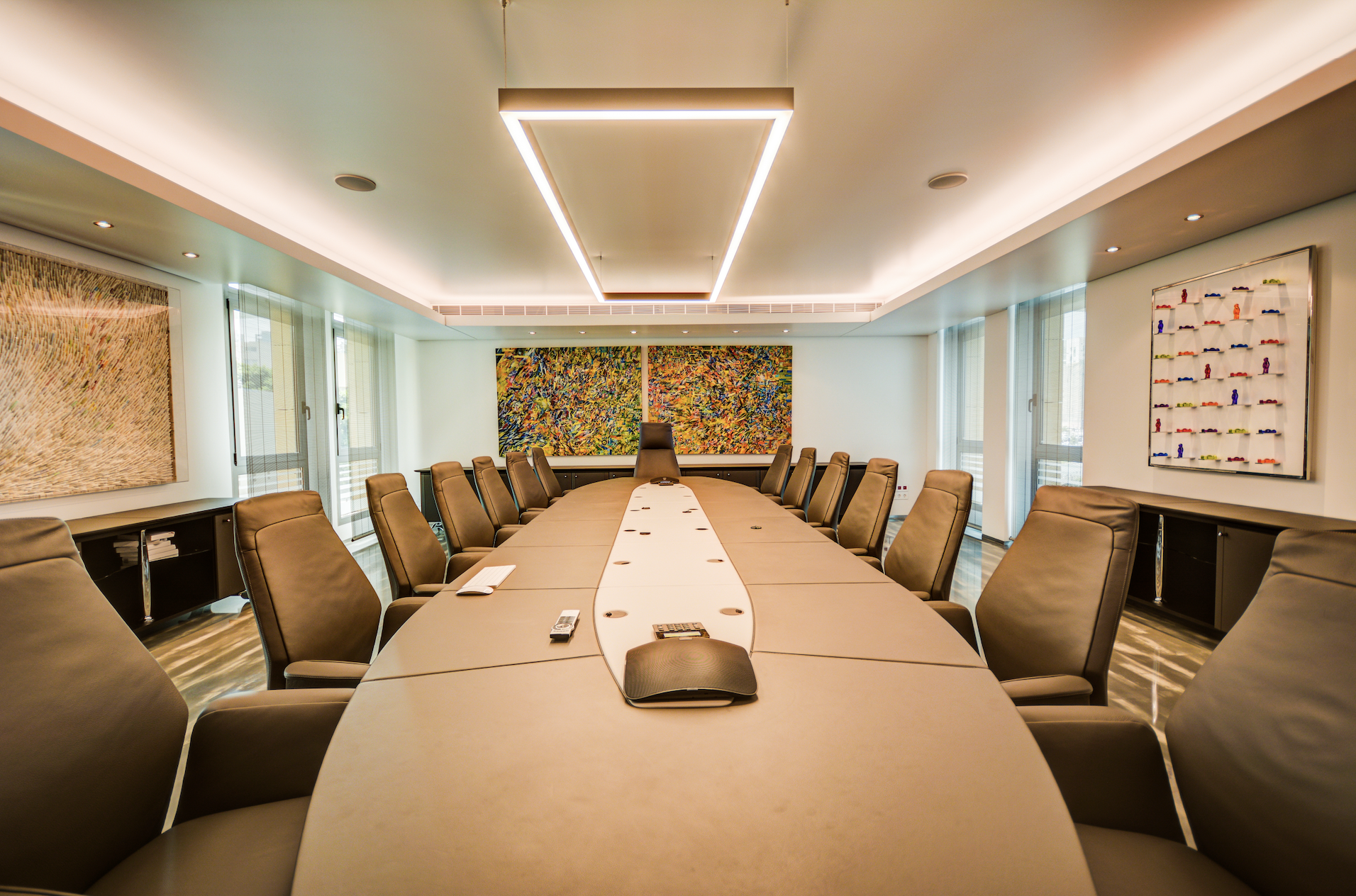 A modern conference room with a large oval table surrounded by leather chairs, featuring abstract artwork on the walls and a sleek ceiling light fixture.