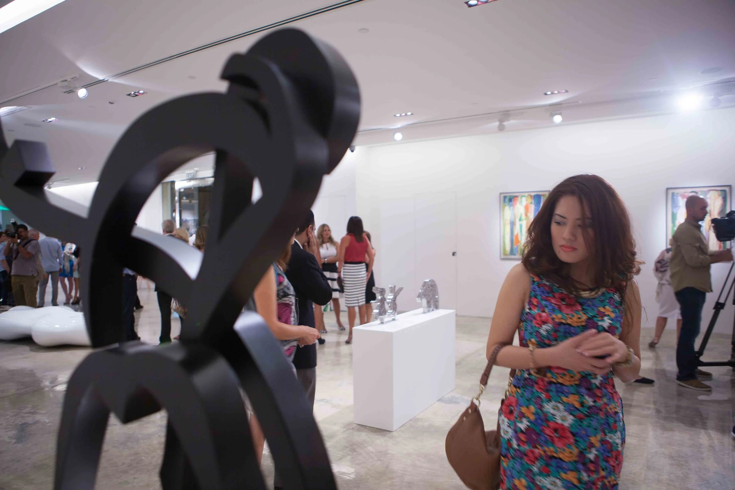 People viewing artwork at an art gallery opening, with a sculpture in the foreground and colorful paintings on the white gallery walls.