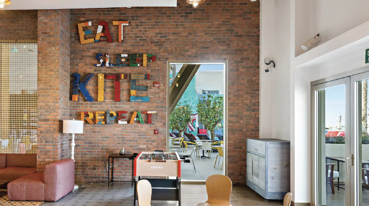 Interior of a colorful, modern room with a brick wall, decorated with large multicolored letters spelling out "EAT SLEEP PLAY REPEAT." There is a foosball table in the center, a pink armchair with a lamp, a small side table, and a cabinet. Through a 
