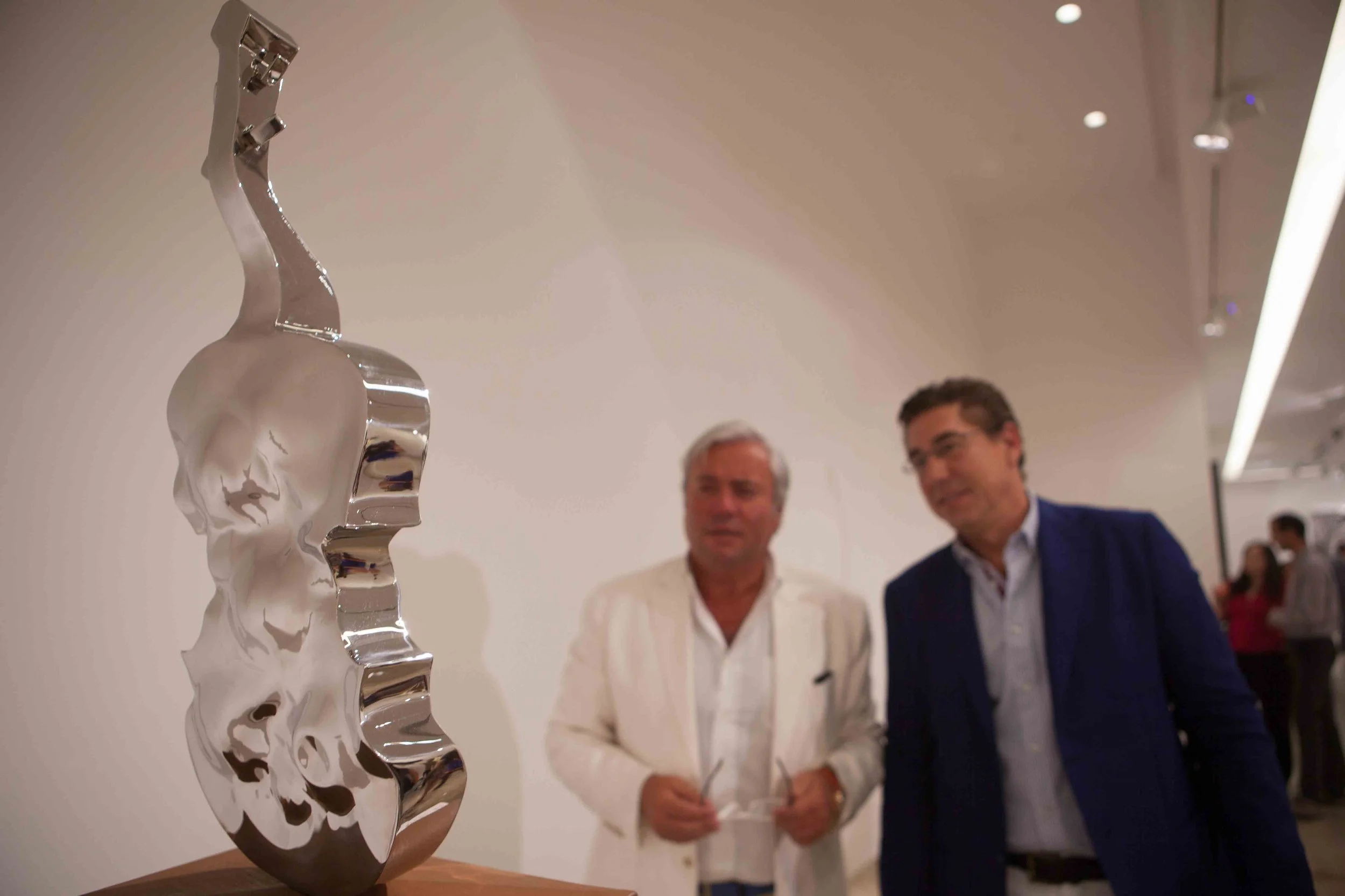 A shiny, abstract metal sculpture on display in an art gallery with two men observing it.