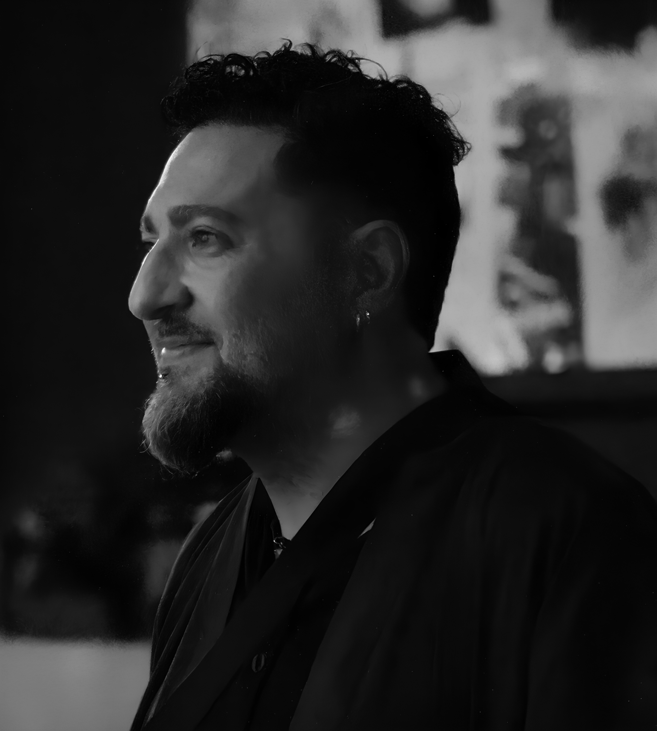 Black and white photo of a man with dark, curly hair and a beard, smiling, wearing a dark jacket.
