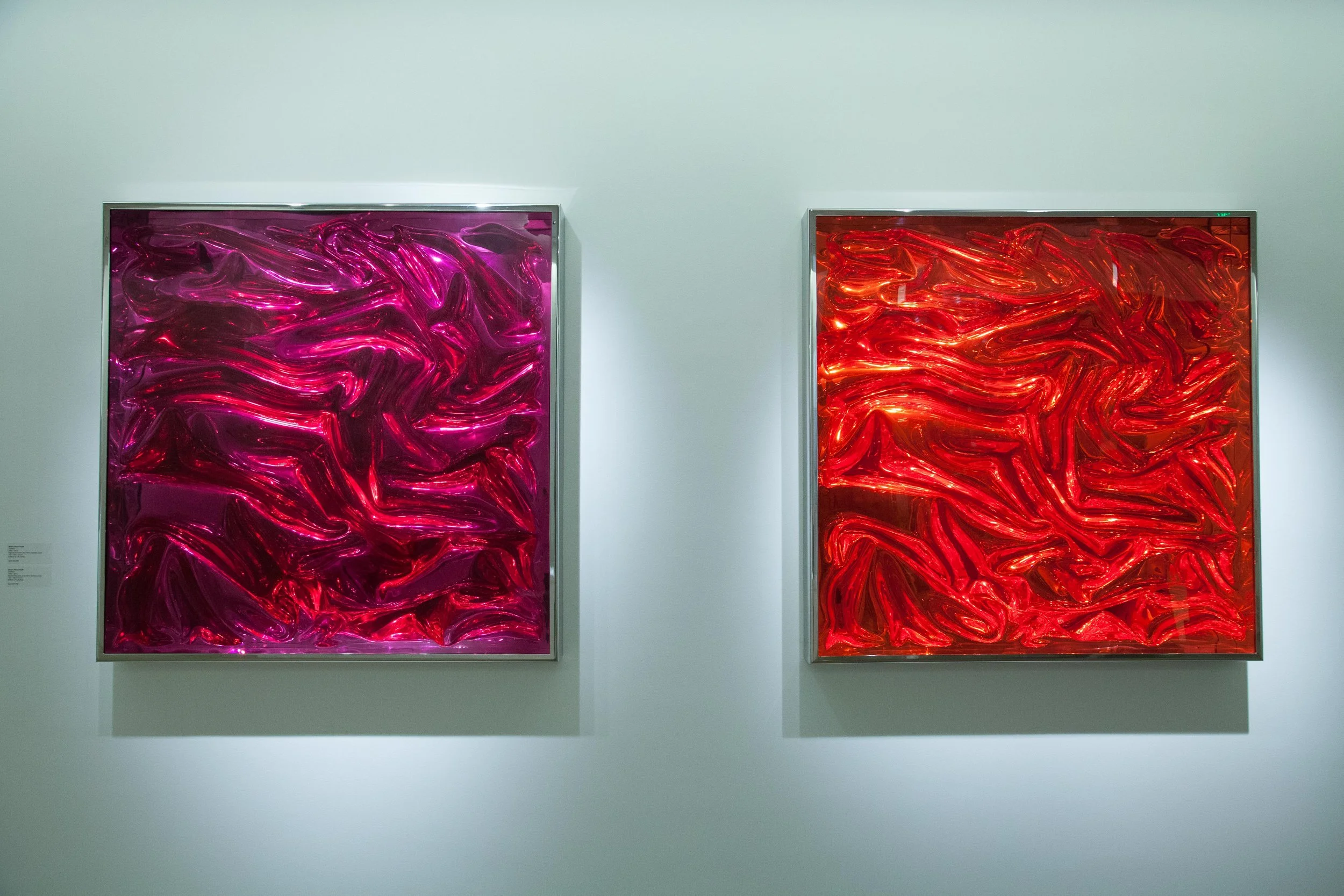 Two abstract glass art pieces with swirling, textured patterns—one pink and purple, the other red—mounted on a white wall in an art gallery.