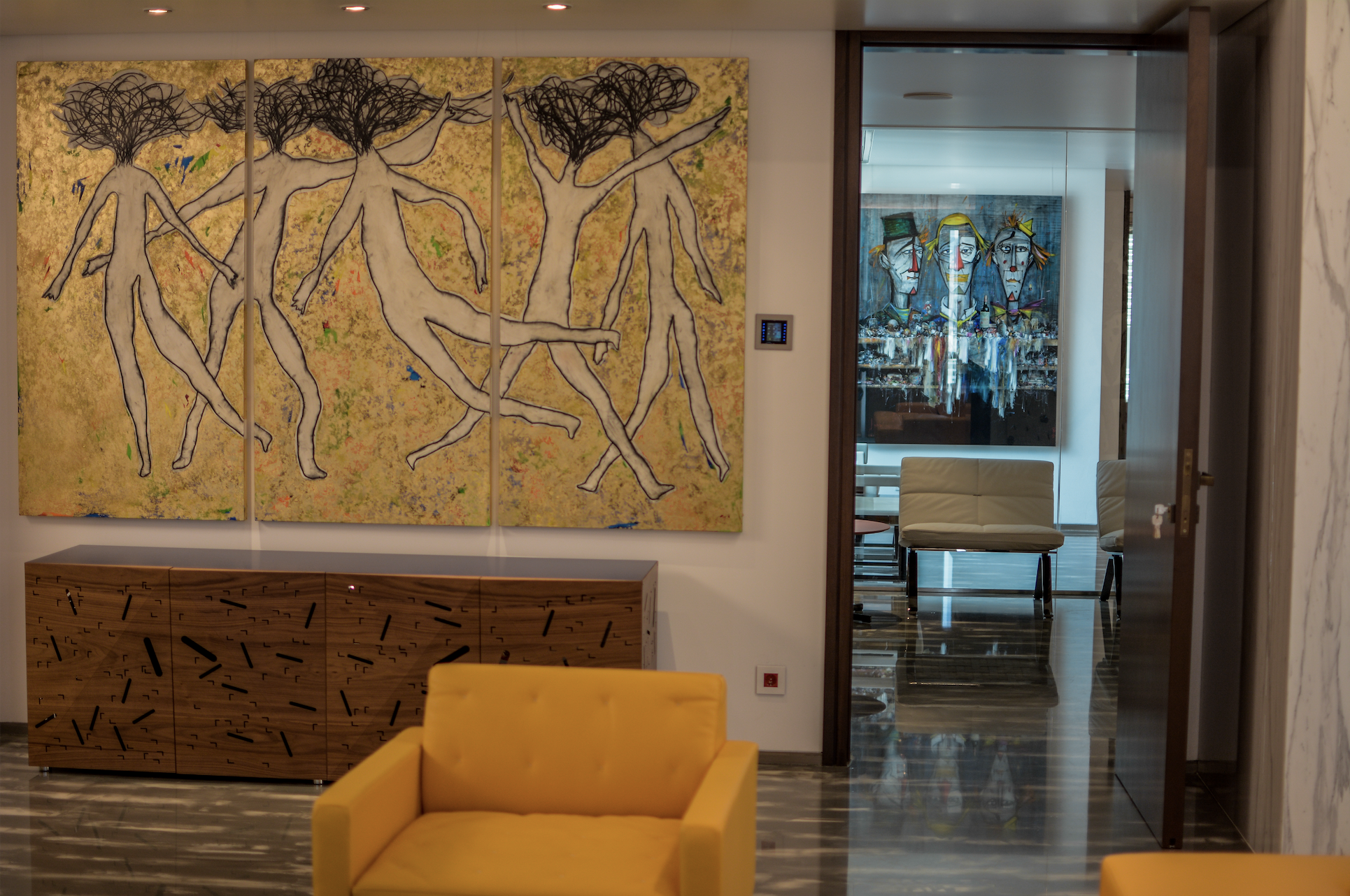 Interior of a modern living room with a yellow armchair, abstract paintings on the walls, and a doorway leading to another room with art, chairs, and a glass table.