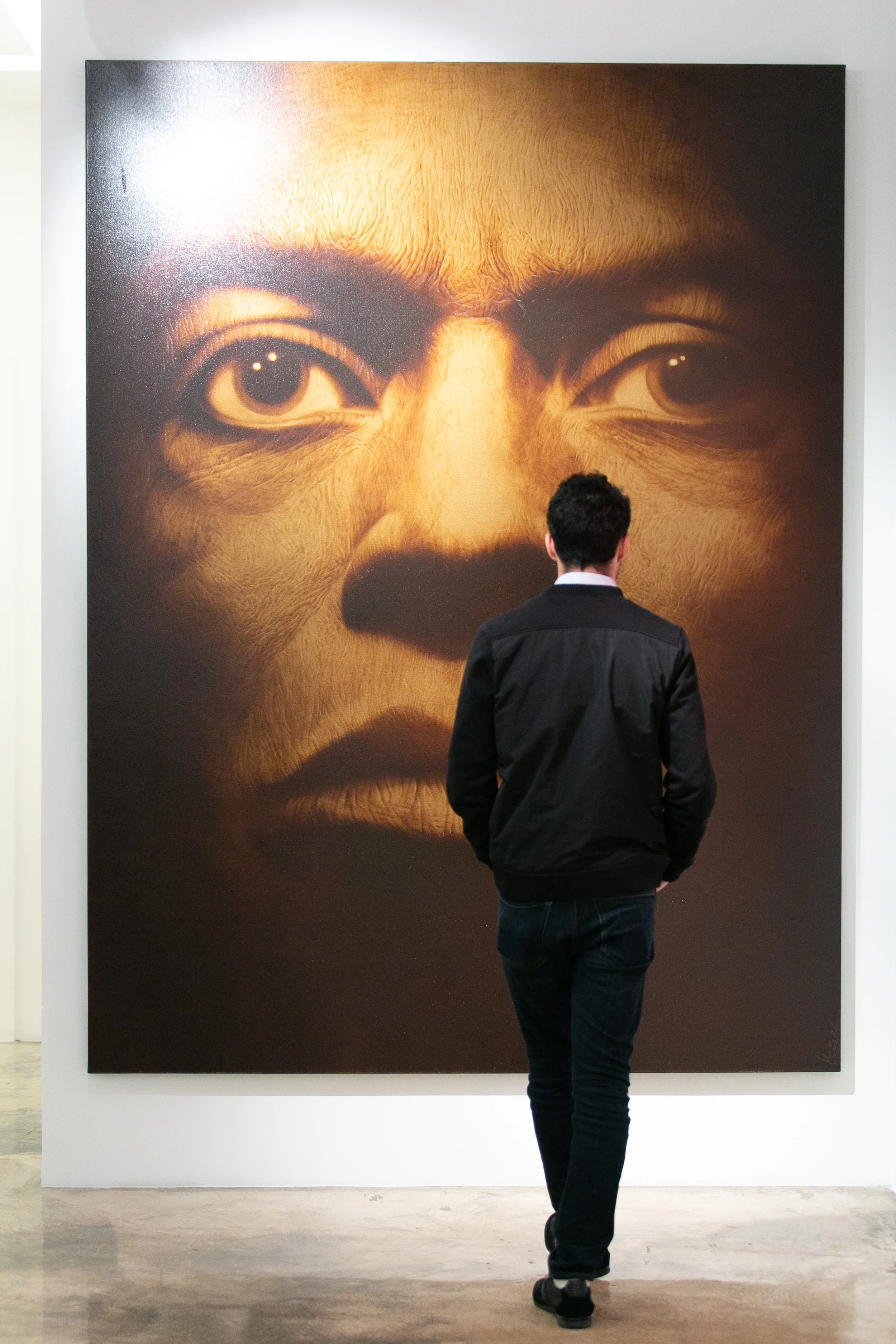 A man with dark hair wearing a black jacket and jeans is looking at a large art piece of a cat's face in an art gallery.
