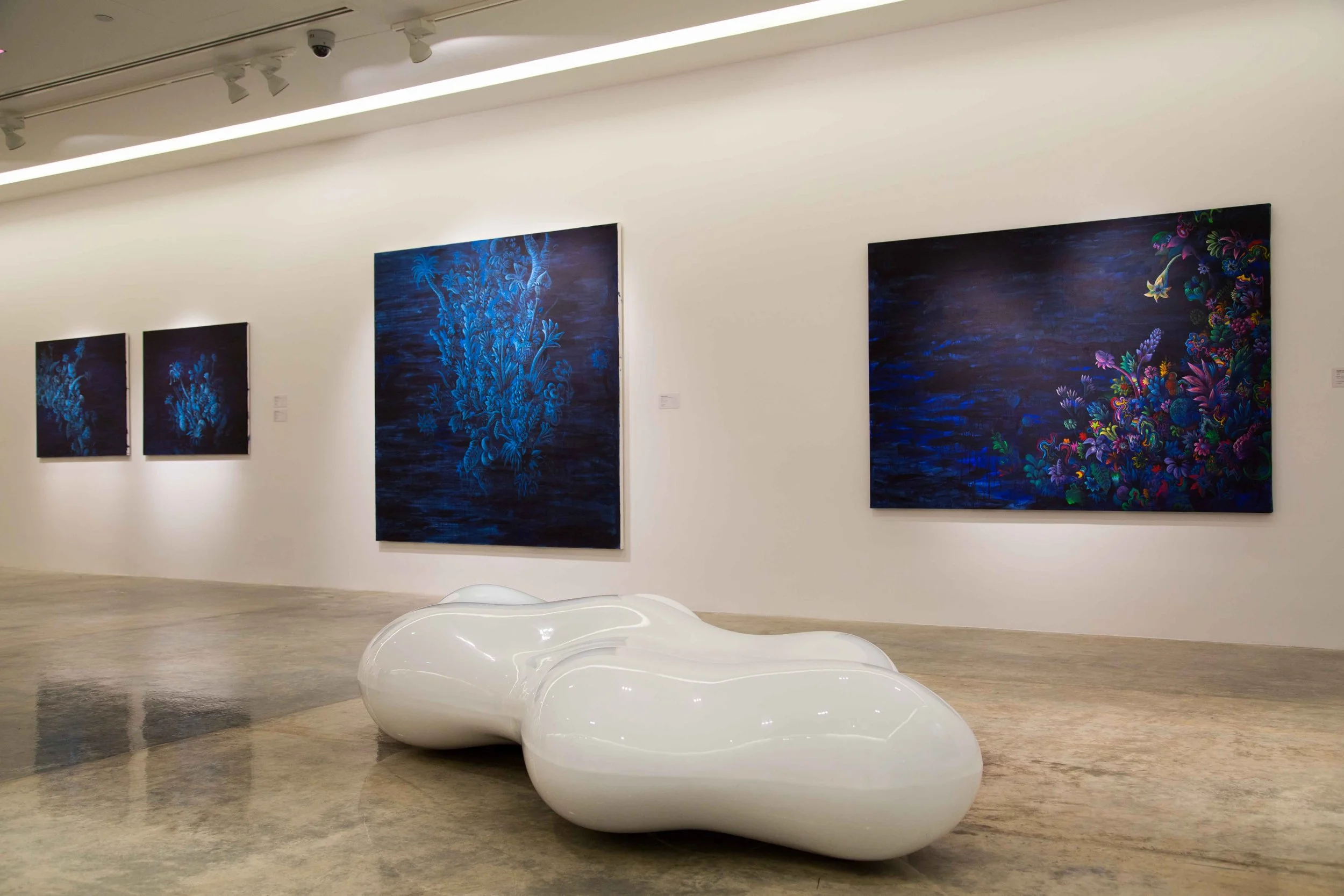 Modern art gallery with three large paintings featuring blue and colorful floral designs on white walls and a white abstract sculpture in the foreground.