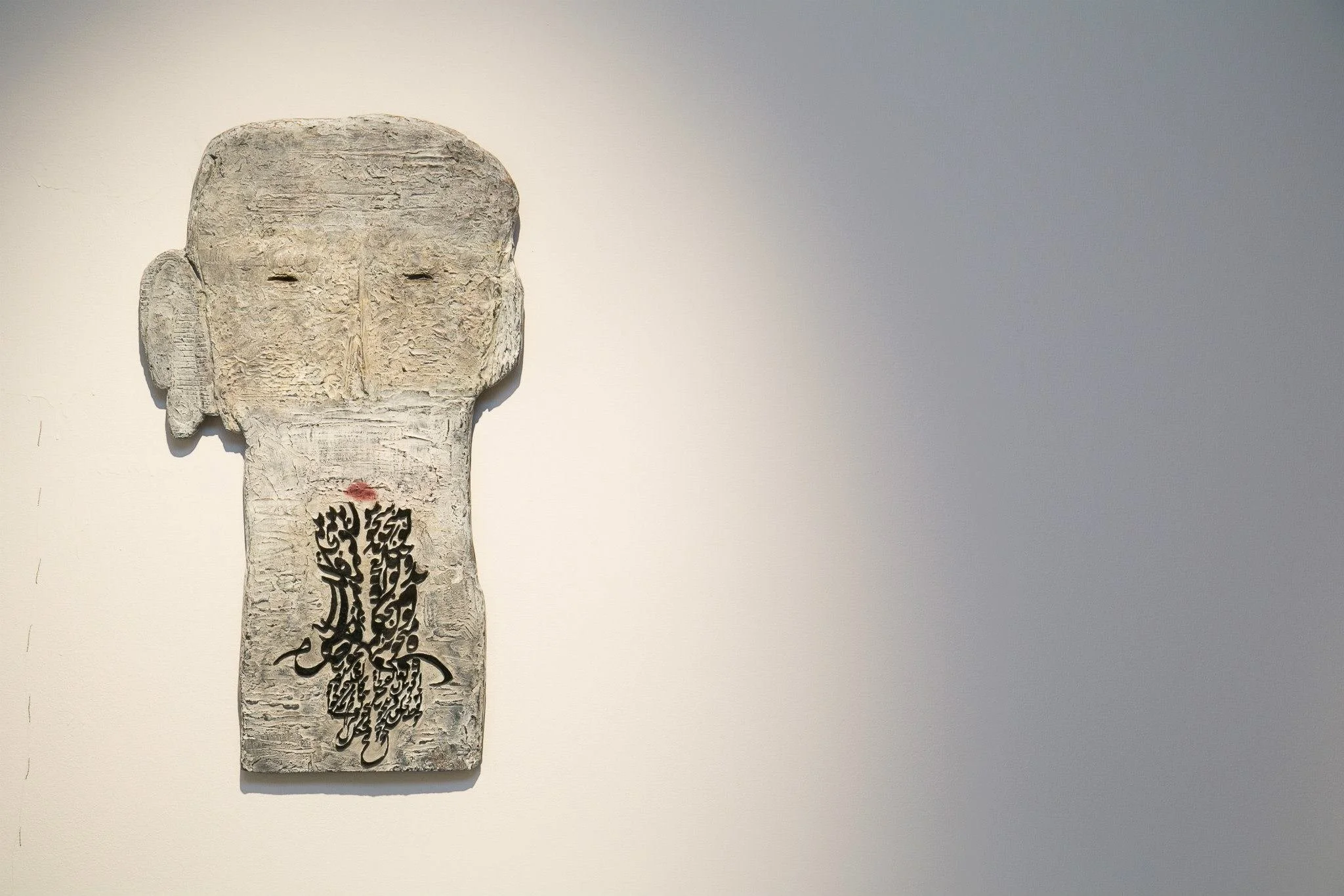 Art sculpture of a human face and upper torso with abstract black calligraphy at the chest, mounted on a white wall.