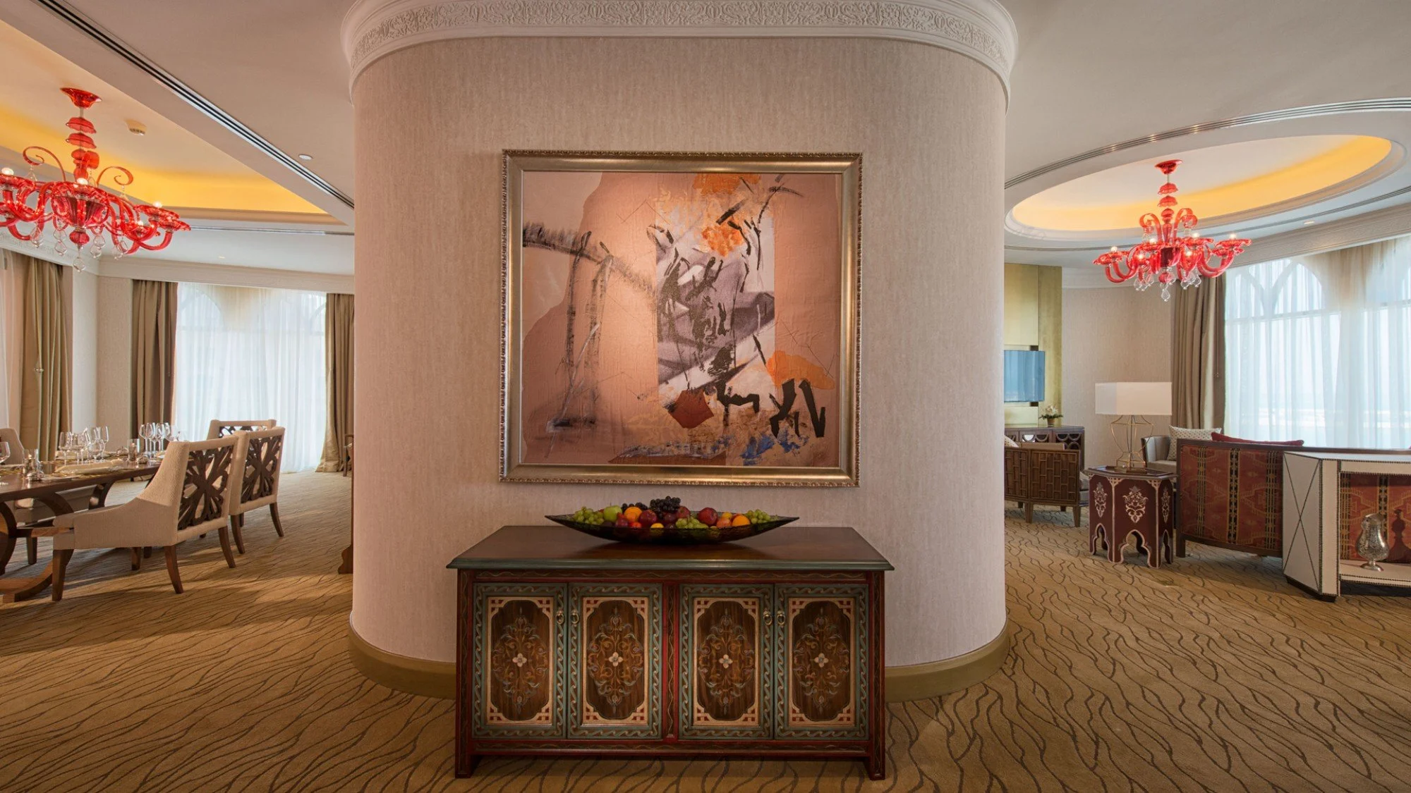 Interior of a luxurious hotel suite with a curved wall featuring a framed abstract painting, a ornate dark wood sideboard with a fruit bowl, and a spacious living area with dining table, chairs, and elegant lighting fixtures.