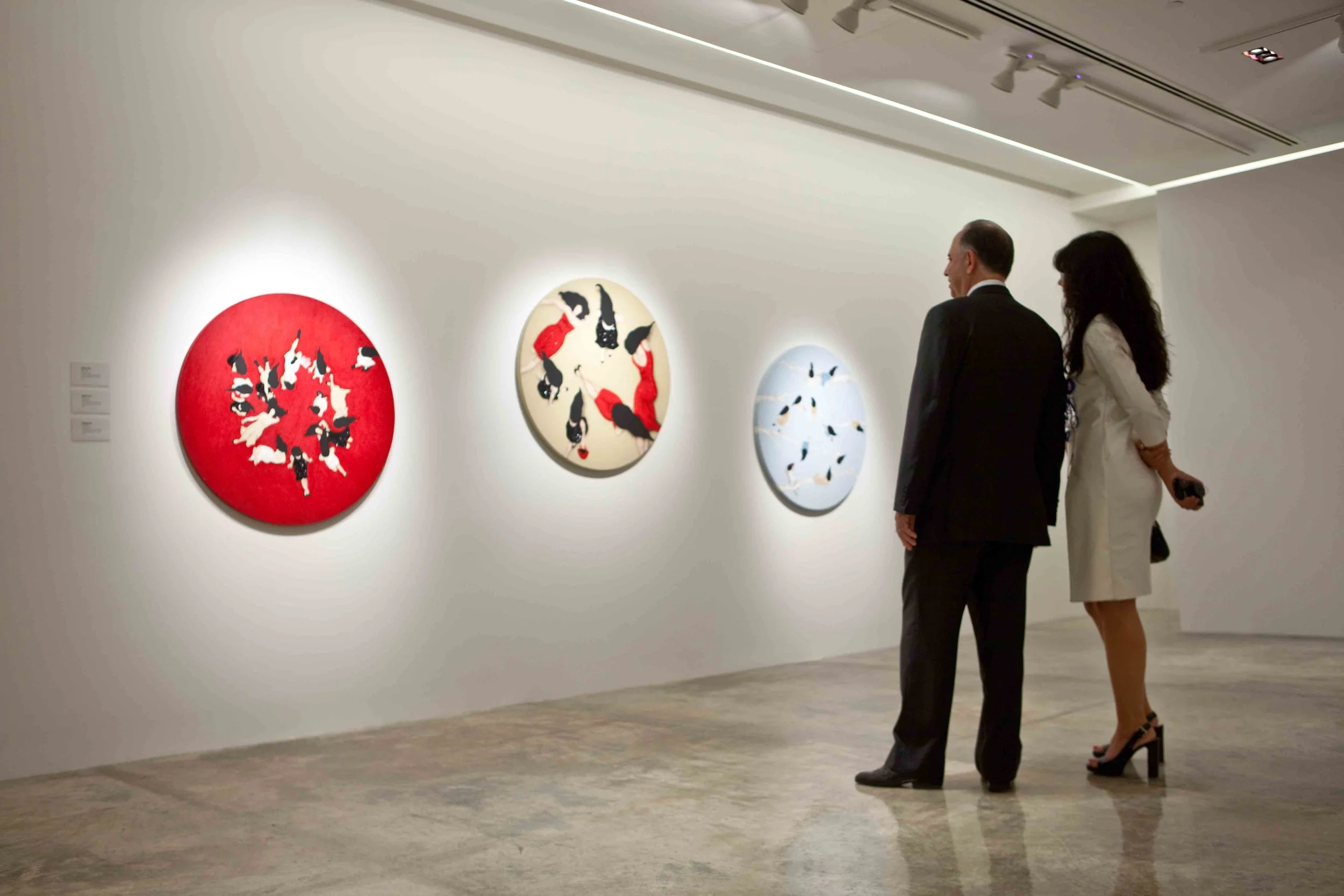 Two people, a man in a suit and a woman in a white dress, are observing abstract art paintings in an art gallery. The paintings are circular, feature various black, white, red, and beige colors, and are hung on a white wall.