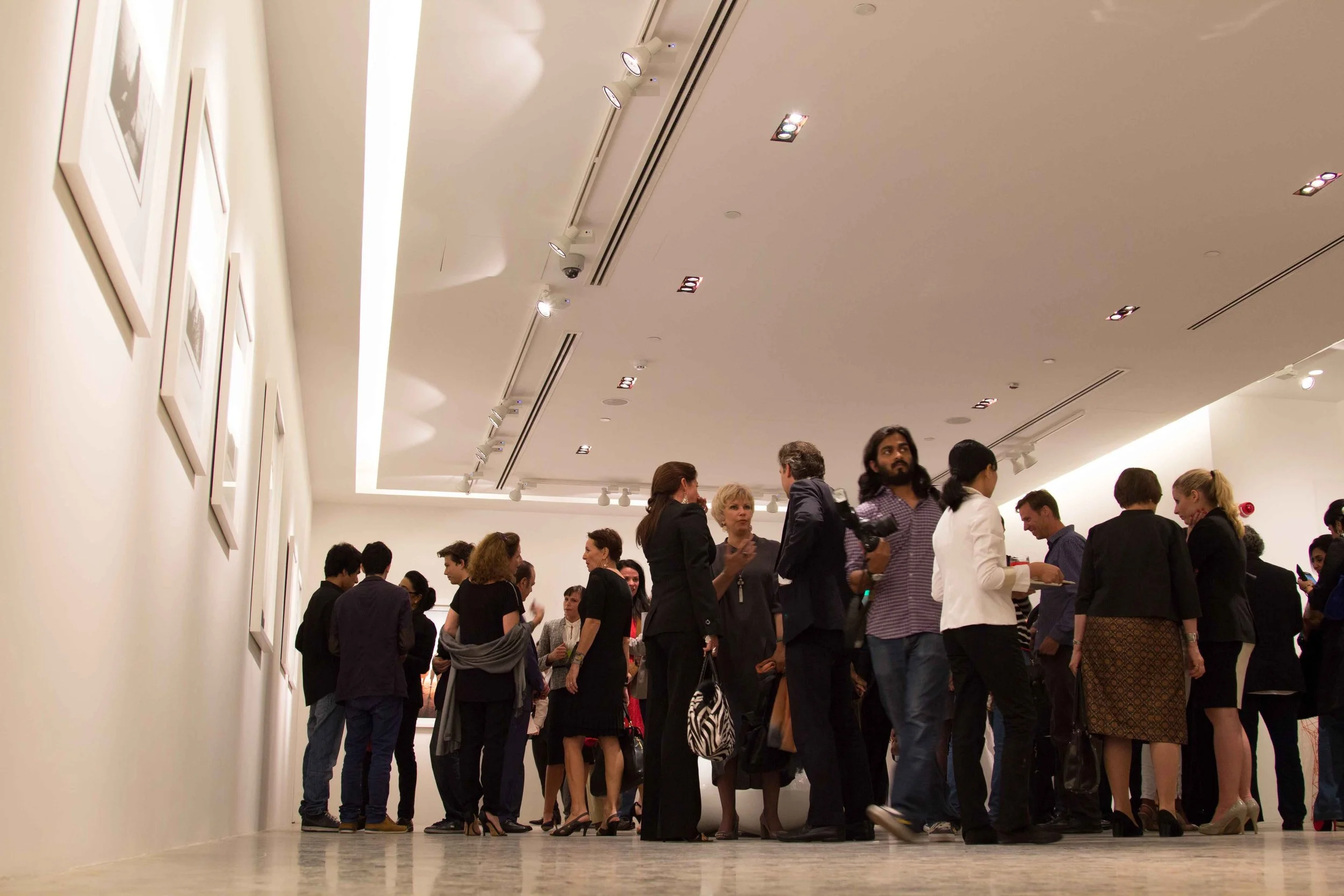 Group of people in an art gallery or museum, standing and talking in a well-lit modern space with framed pictures on the wall.