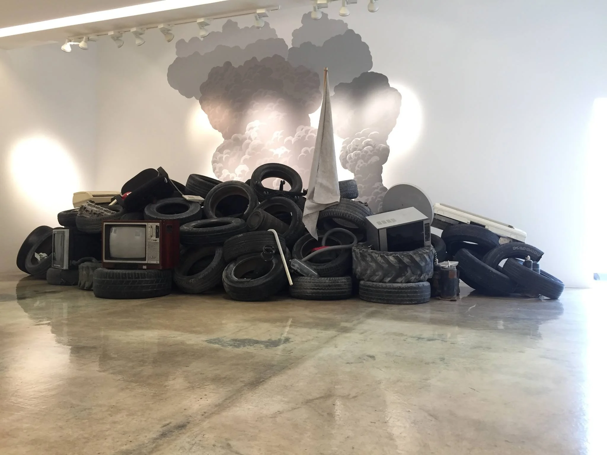 A pile of discarded tires and old electronic appliances, including an TV and microwave, is in an art installation with a background mural of cloud-shaped smoke.