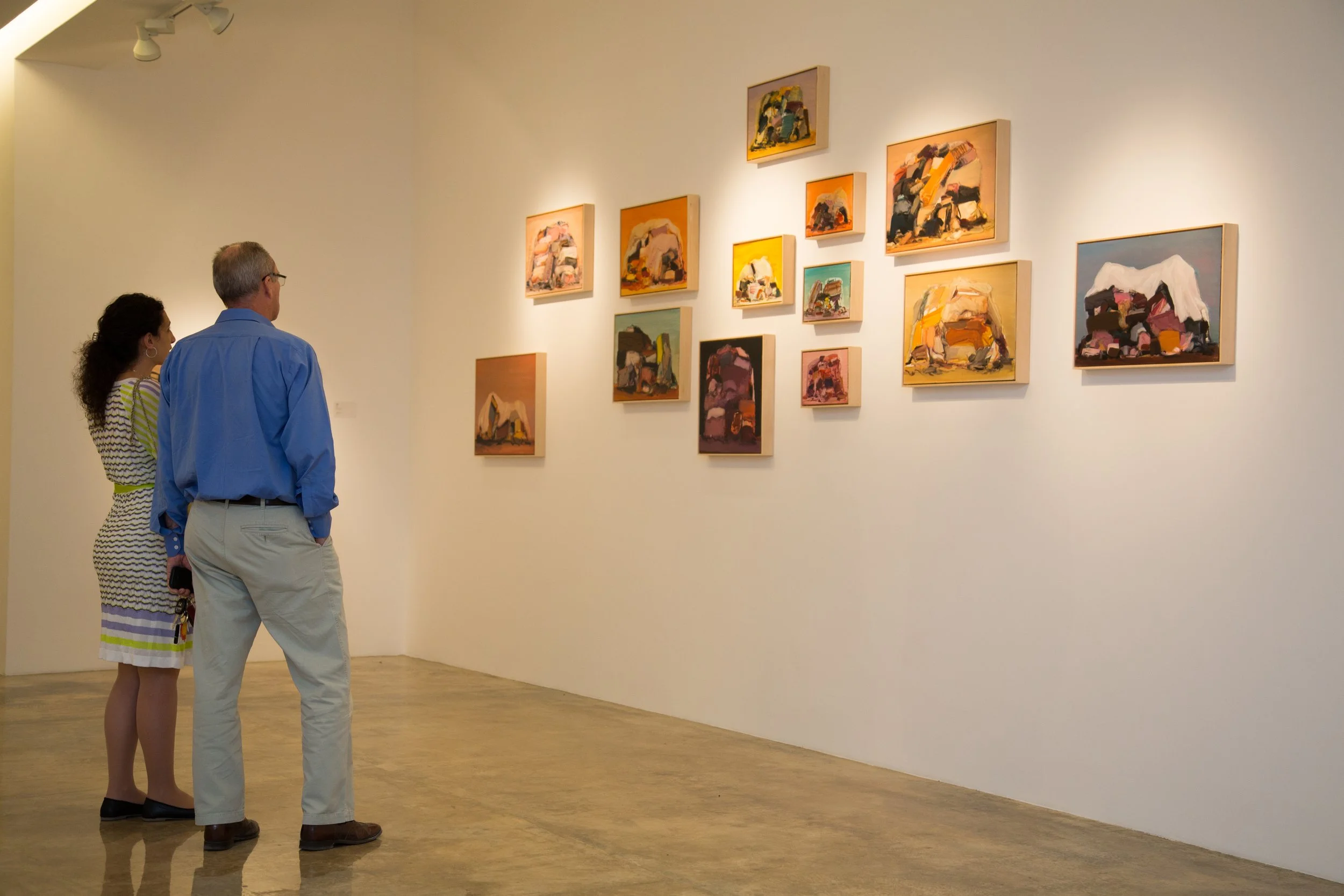 Two people, a woman with curly hair in a patterned dress and a man with glasses in a blue shirt, viewing abstract paintings in an art gallery with white walls and wooden floors.