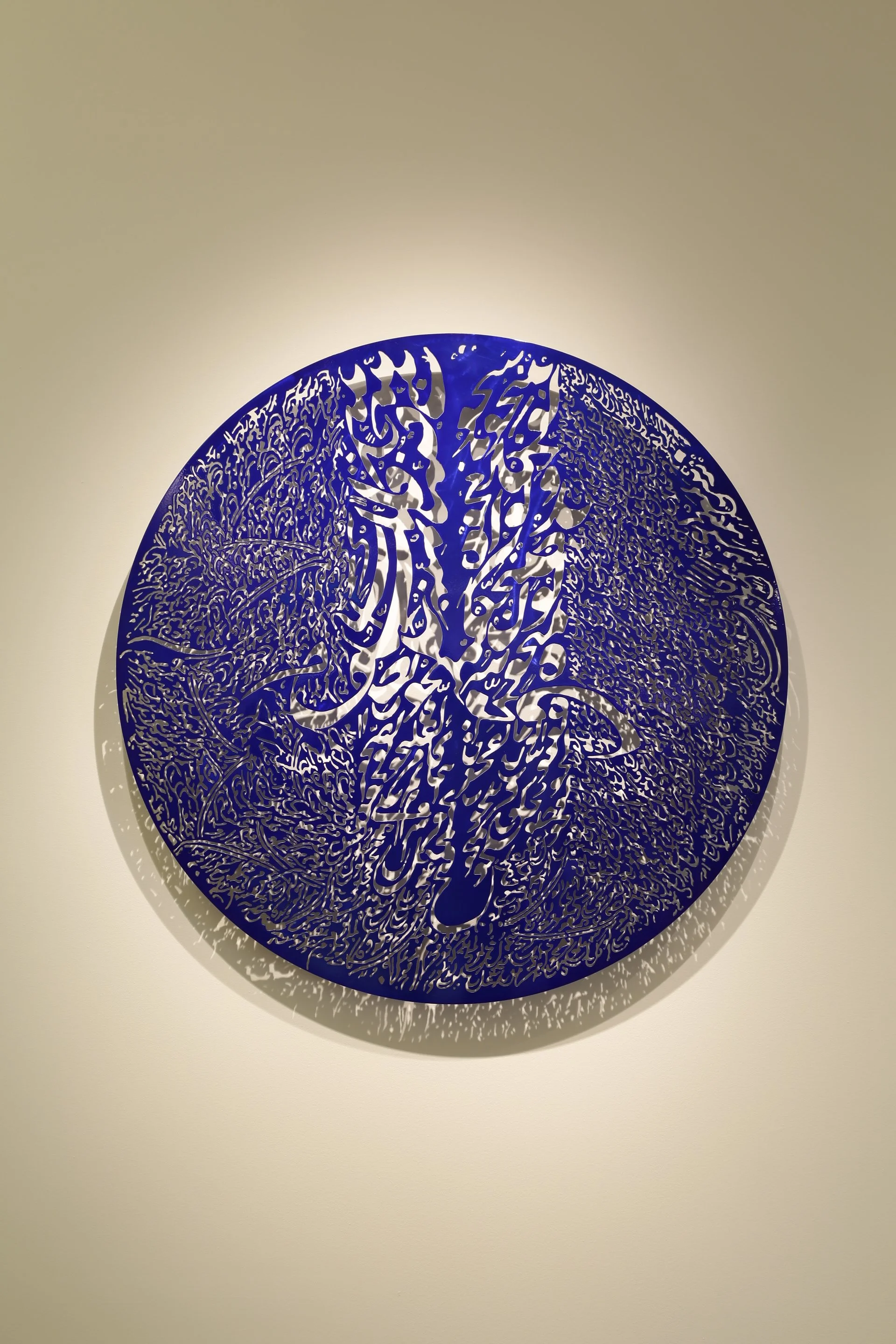 A circular decorative art piece with intricate cut-out Arabic calligraphy in blue, casting a shadow on a plain wall.