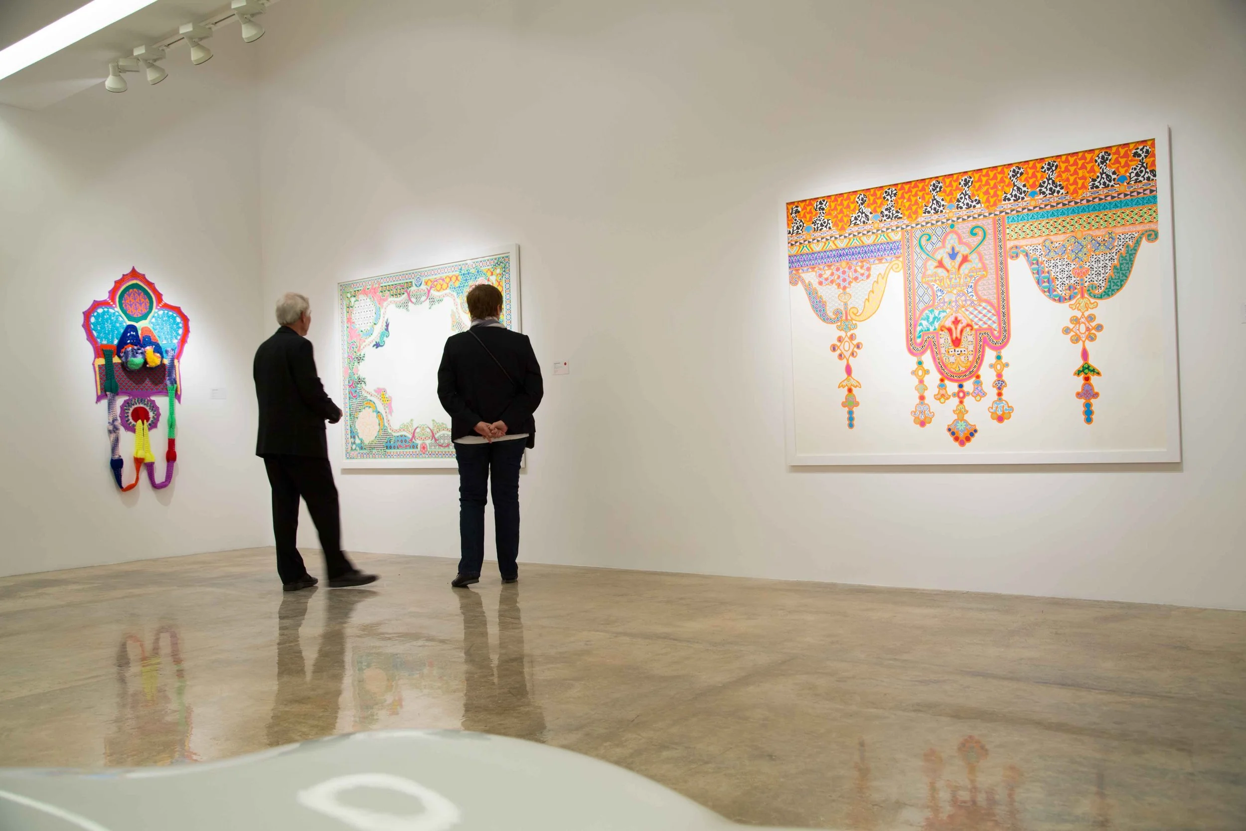 Two people viewing colorful modern artwork on white gallery walls. The artwork includes a hanging textile piece and two framed paintings with intricate patterns and vibrant colors.