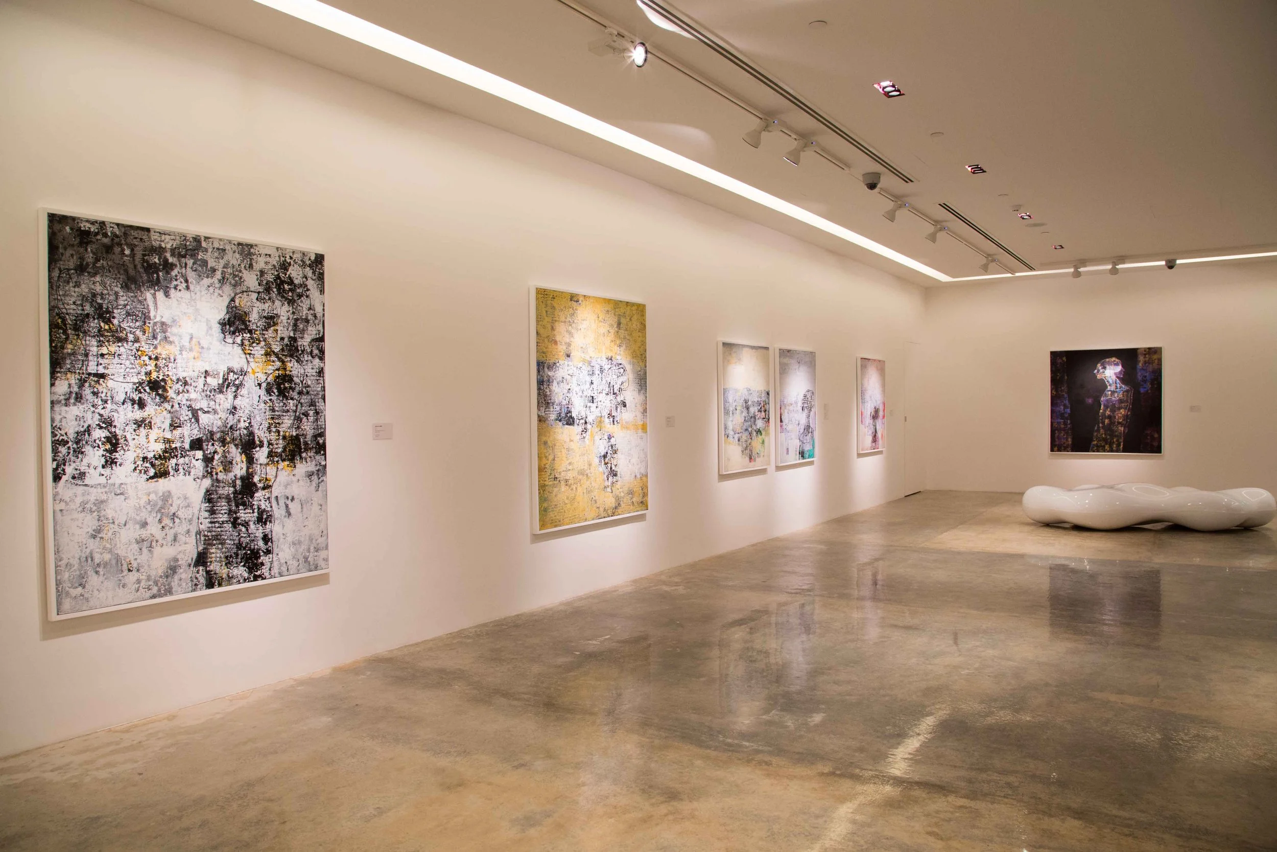 Art gallery with several abstract paintings on white walls and a modern white sculptural bench on a polished concrete floor.