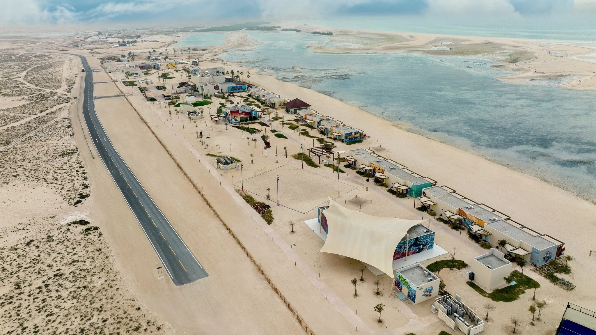 Aerial view of a beachside amusement park with colorful structures, boardwalks, pathways, and a large stage, along a sandy shoreline with a calm water body in the background and a road running parallel to the beach.