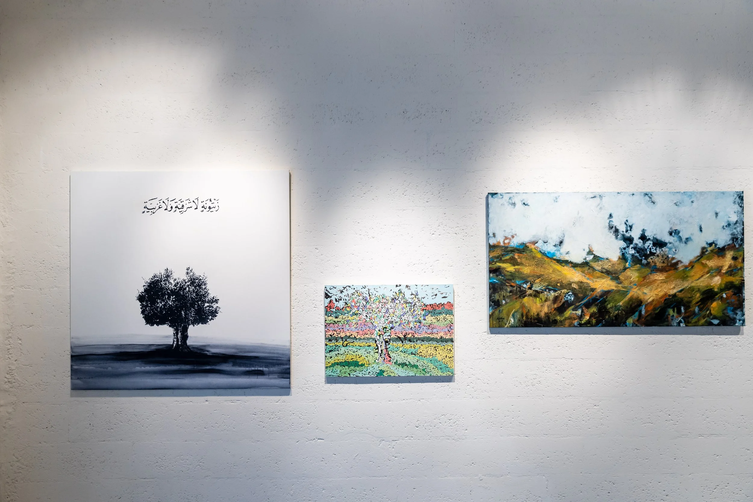 Three paintings on a white gallery wall: a black and white drawing of a tree with Arabic script above it on the left, a colorful abstract landscape in the middle, and a vibrant mountain scene to the right.
