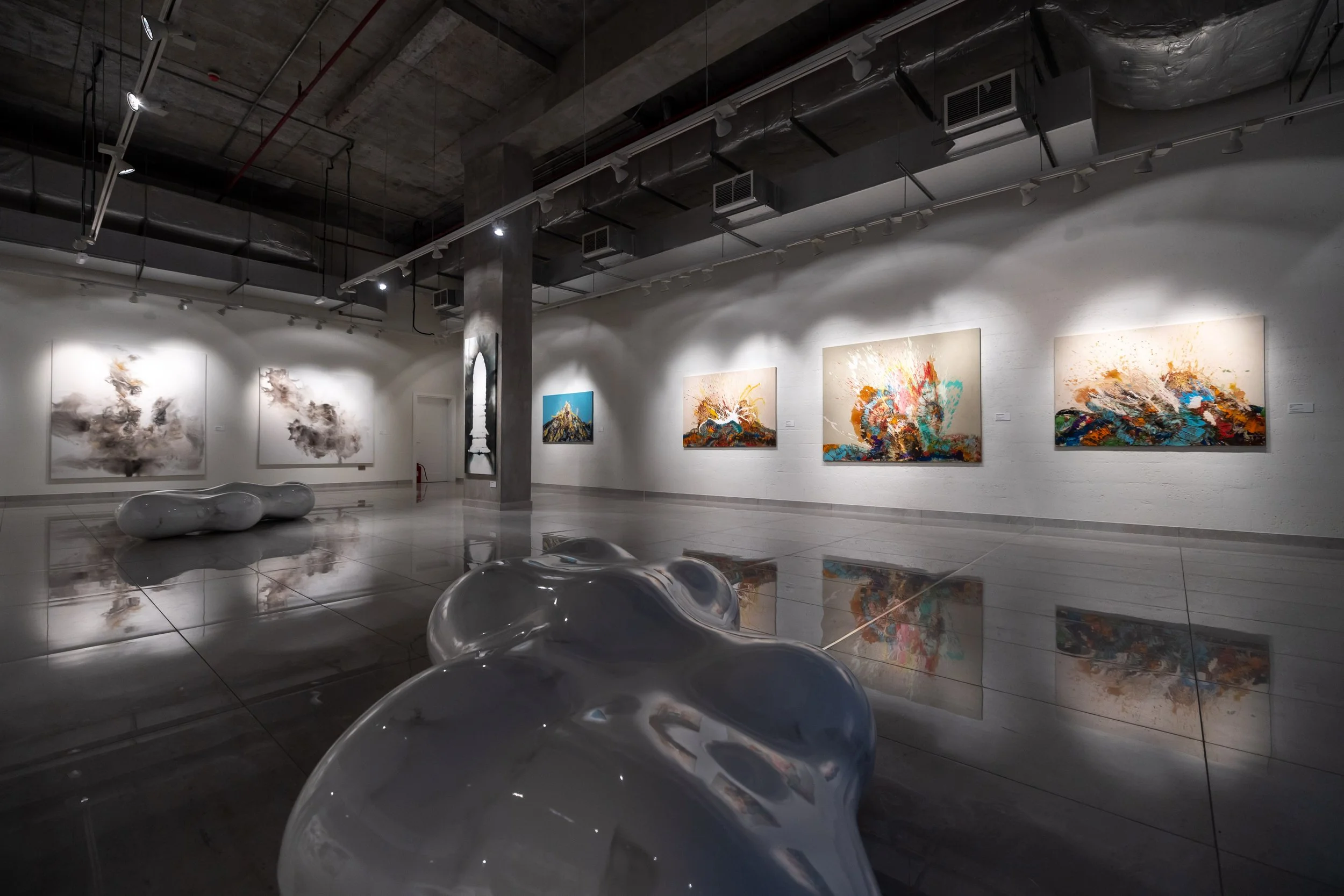 Contemporary art gallery with abstract paintings on white walls and sculpture pieces on the glossy floor, with industrial ceiling and track lighting.
