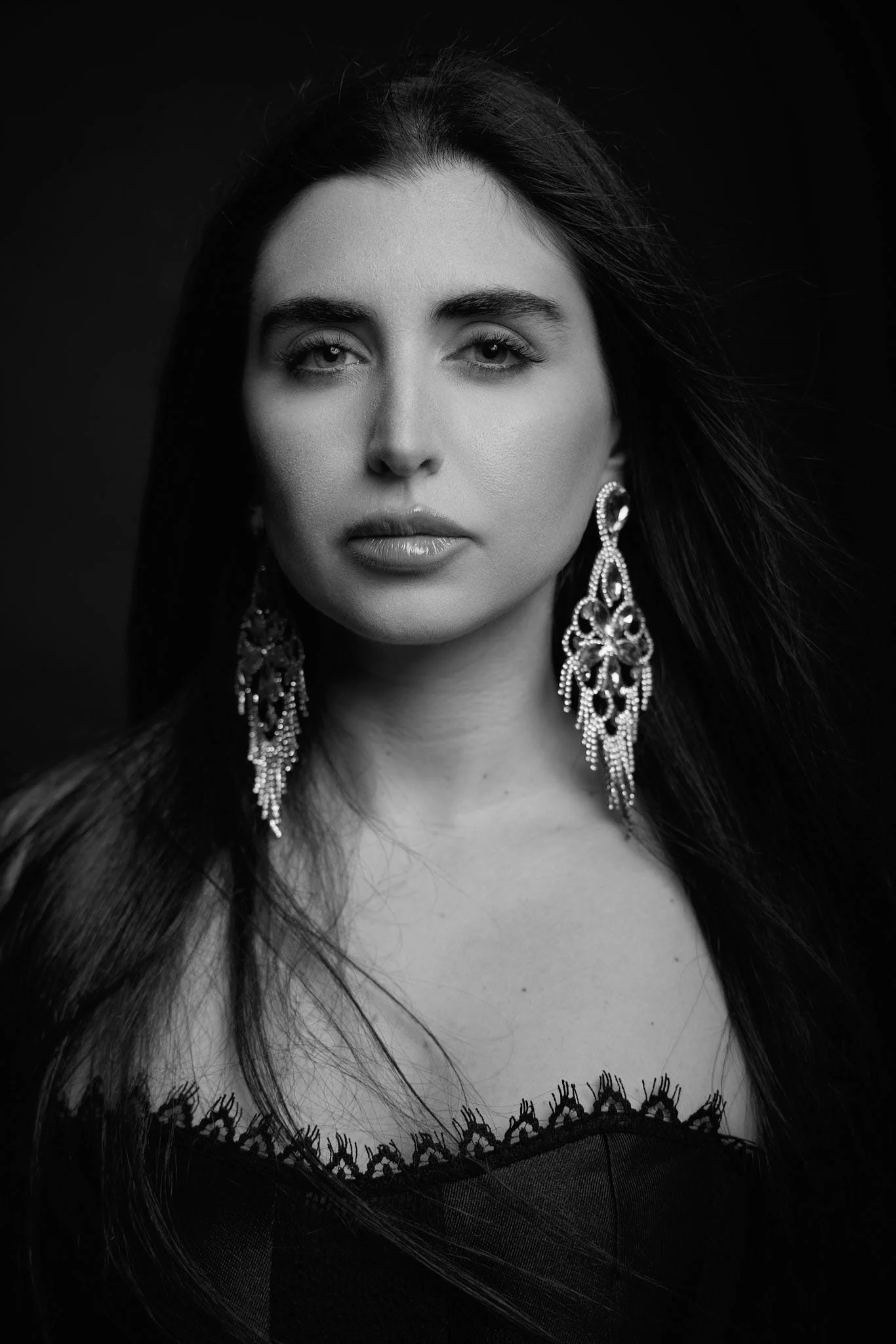 Black and white portrait of a woman with long dark hair, wearing large ornate earrings and a dress with lace detailing at the neckline.