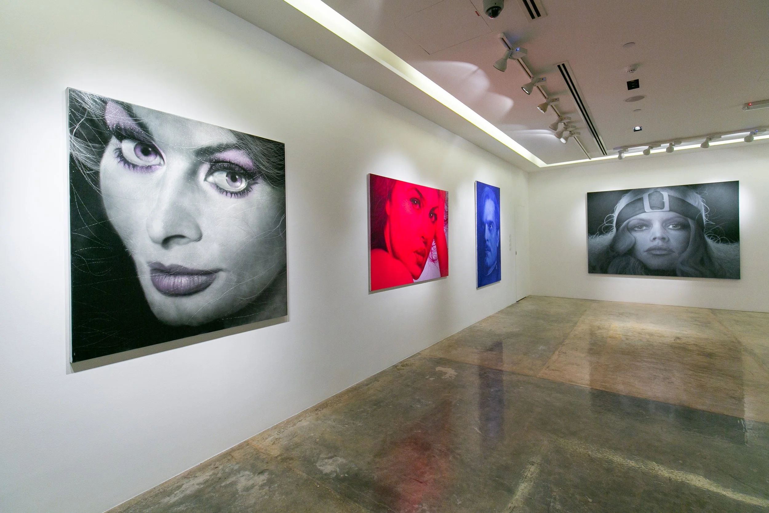 Art gallery with four portraits on white walls, two in color and two in black and white, each depicting women with different expressions and accessories, under spotlight track lighting.