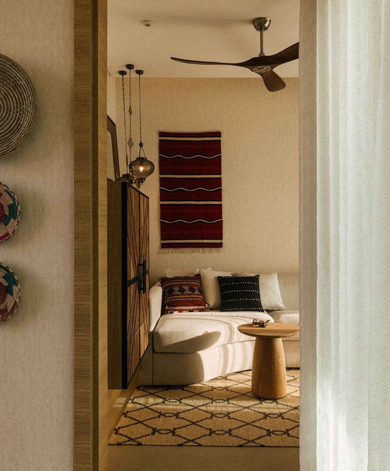 Living room with beige walls, a white sofa with decorative pillows, a small wooden round table, a patterned rug, wall art, a ceiling fan, and hanging pendant lights.