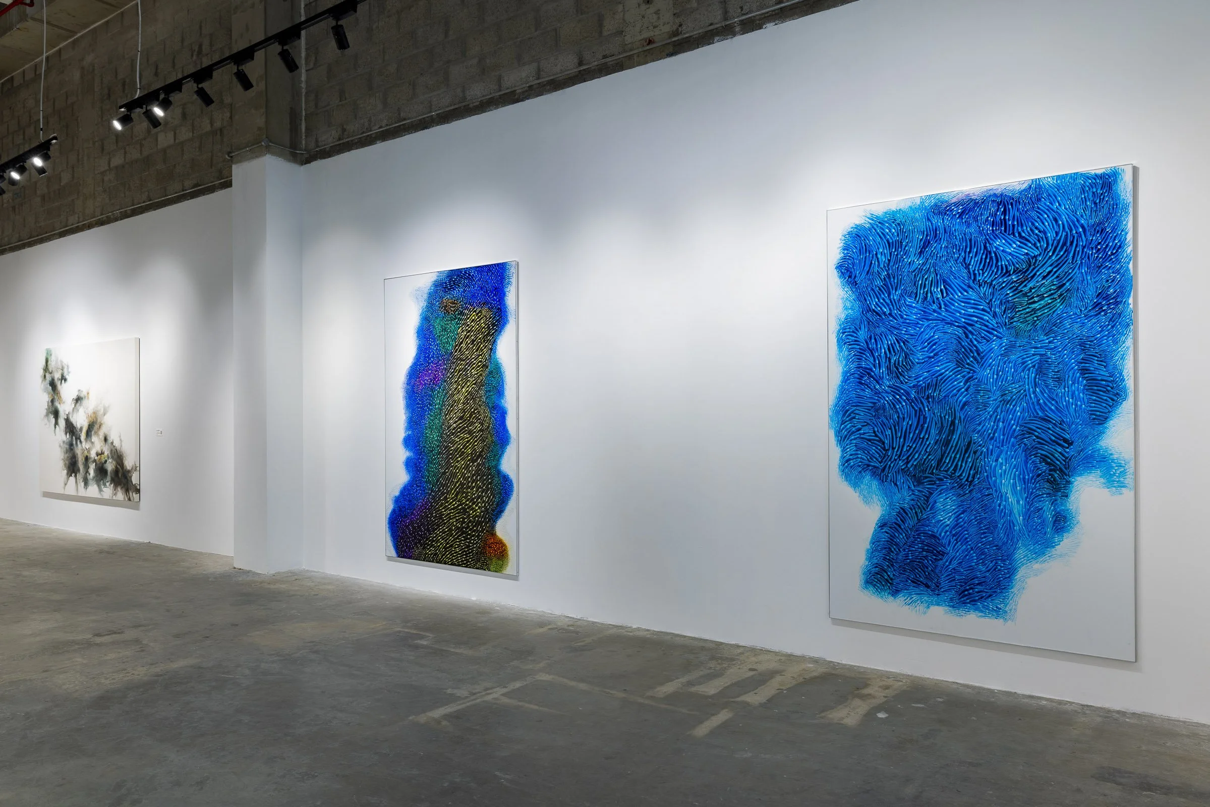 Art gallery with three large abstract paintings on white walls, illuminated by ceiling track lights, in an industrial-style space with concrete floor and exposed brick ceiling.
