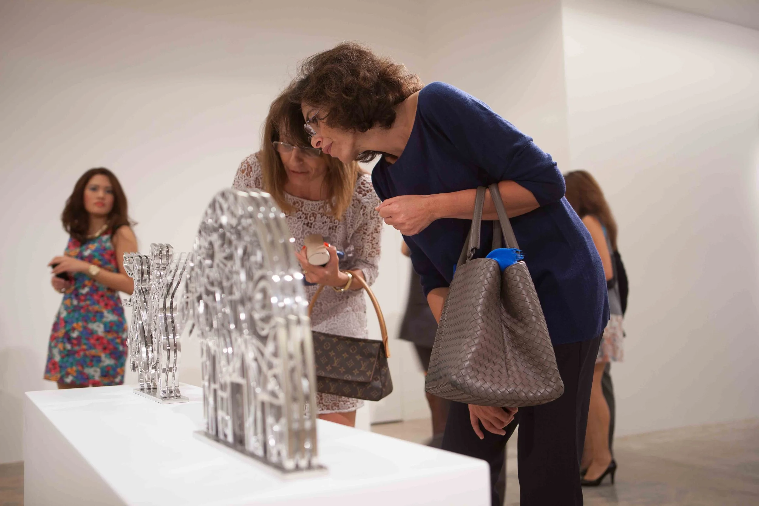 Two women closely examining metallic sculptures on display in an art gallery, with other visitors in the background.