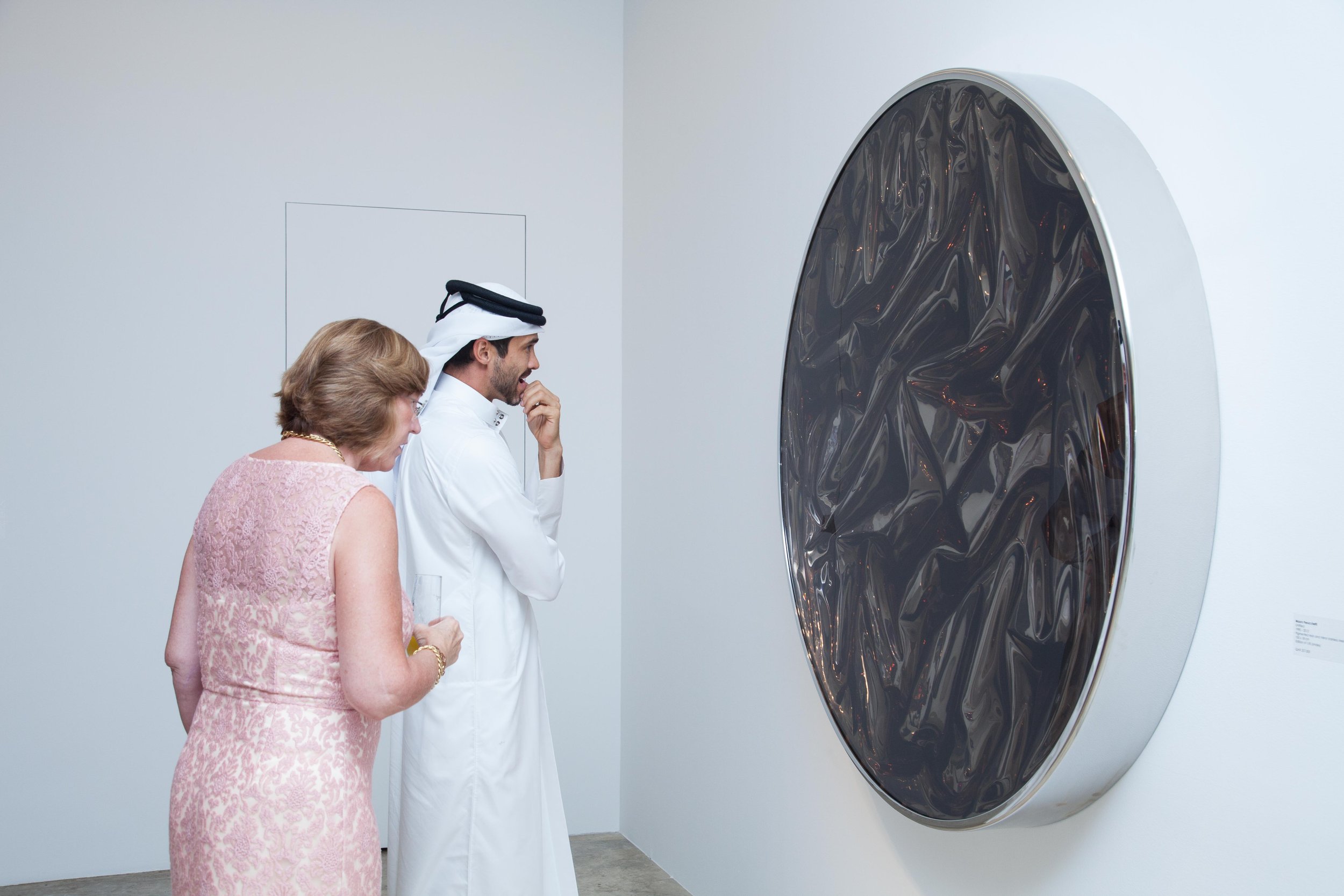 Two women and a man in traditional Middle Eastern attire observing contemporary art in a gallery, featuring a large circular mirrored sculpture on a white wall.