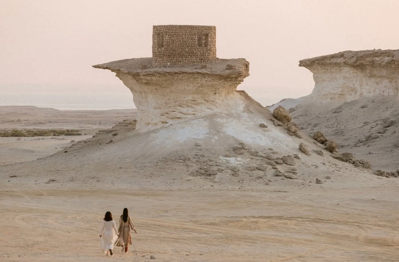 Two women in dresses walking in a desert with rocky formations and a unique stone structure on top of a large outcrop.