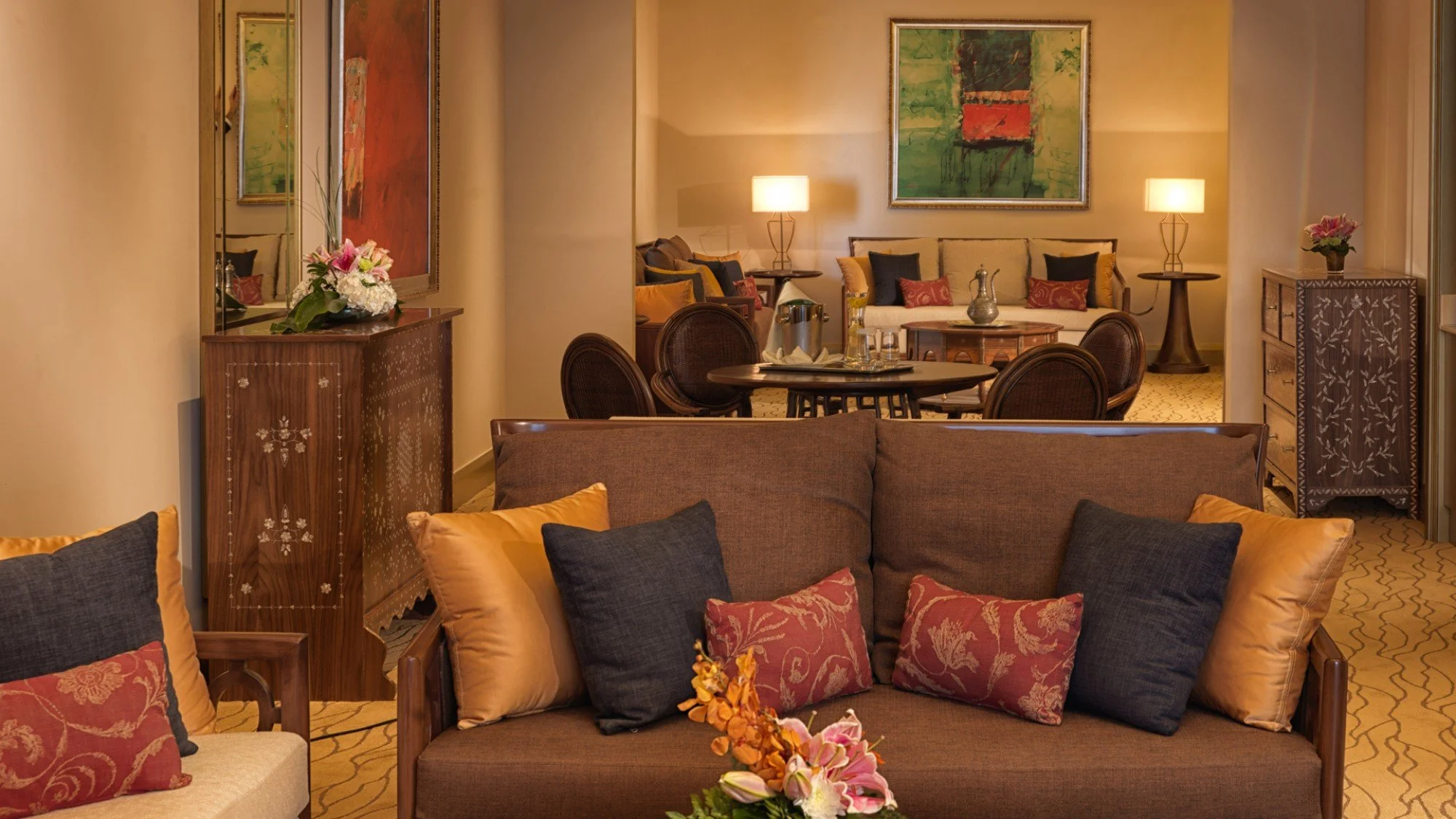 Elegant hotel living room with couches, armchairs, and floral decorations, featuring warm lighting, colorful abstract paintings, and wooden furniture.
