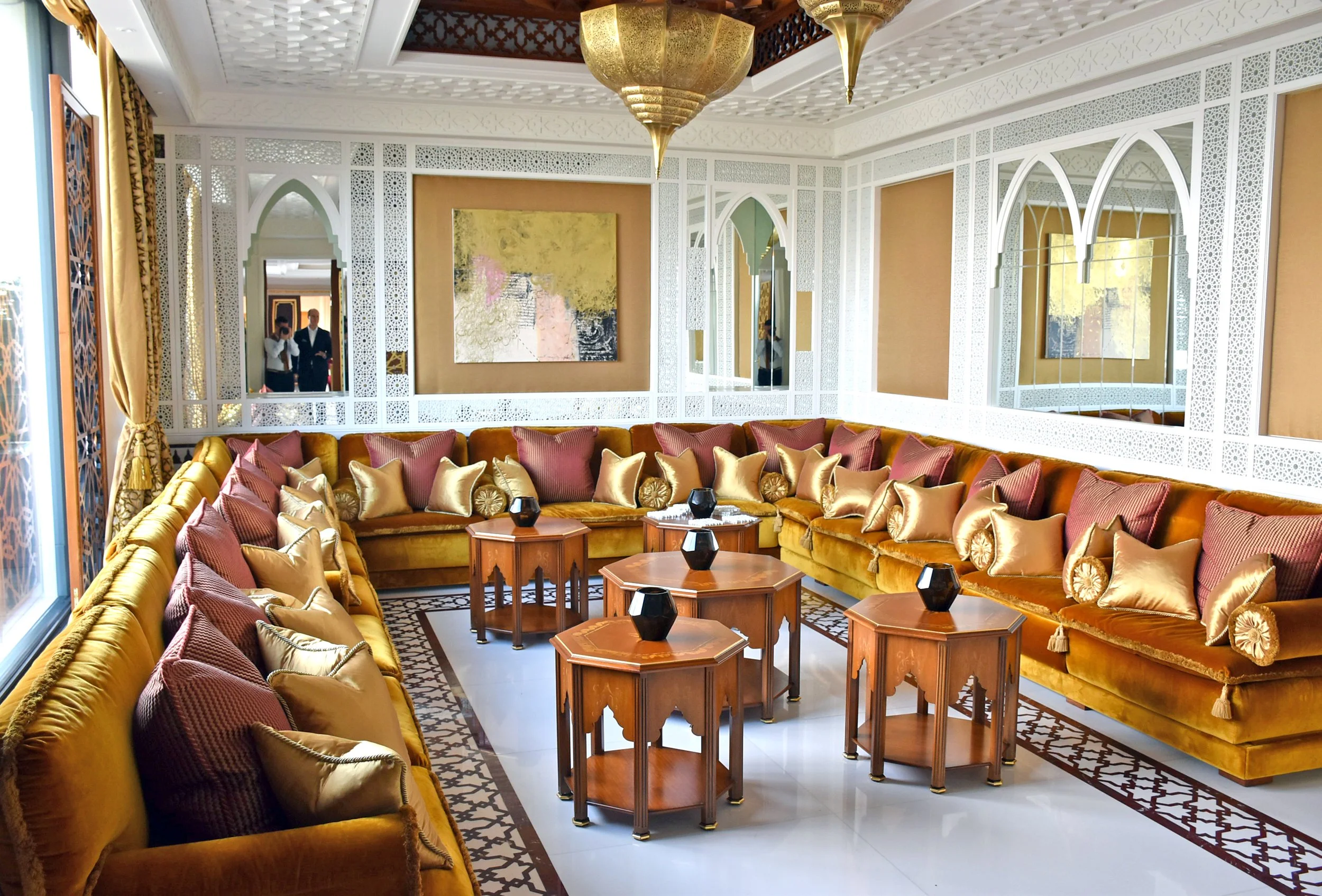 A luxurious living room with a large, U-shaped mustard velvet sofa adorned with pink and gold pillows. In front of the sofa, there are five wooden hexagonal tables with black vases on top. The walls feature intricate white lattice designs, arched mir