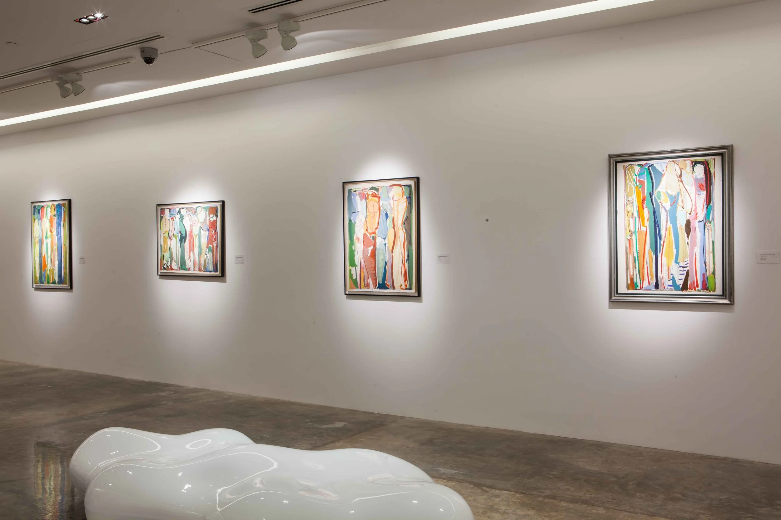 An art gallery with four colorful abstract paintings on a white wall and a white sculptural bench in front.