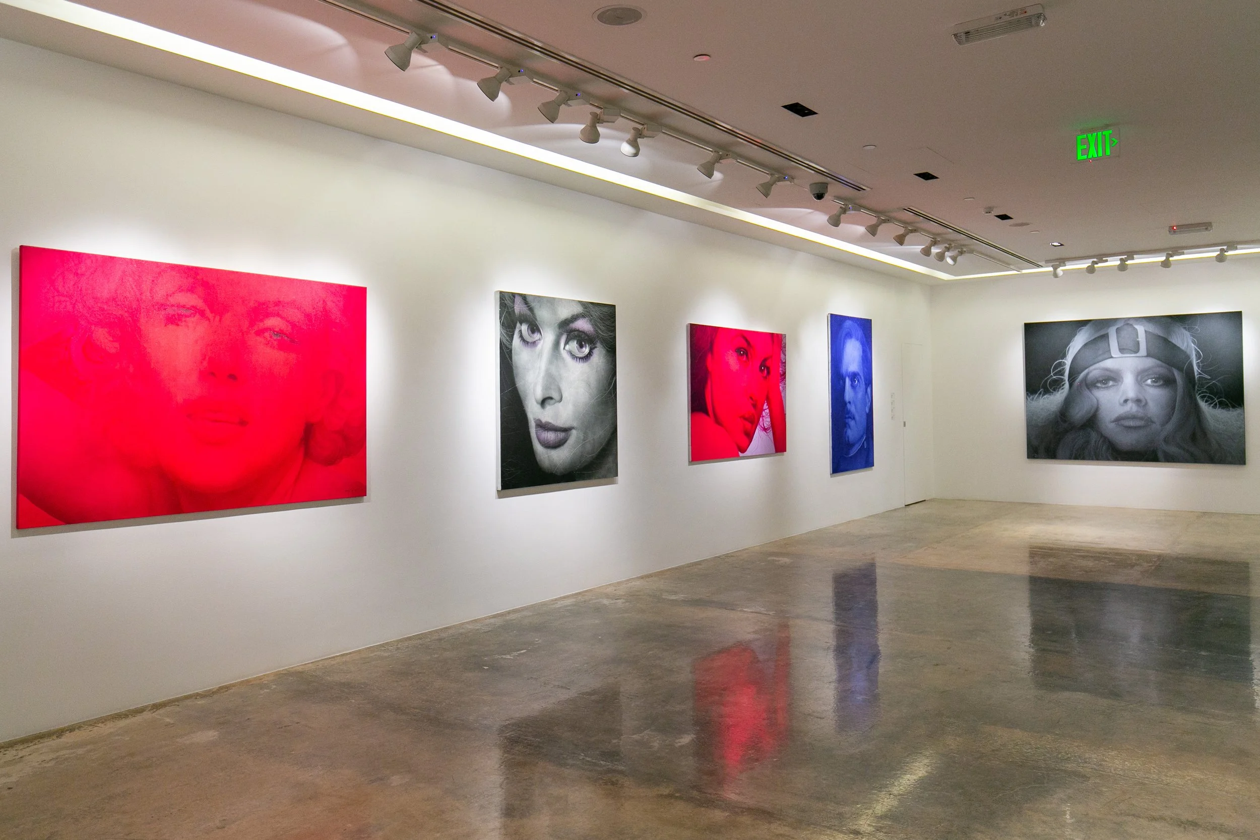 Gallery wall displaying five portrait photographs of women in various colors with different expressions, mounted on white walls inside an art gallery.