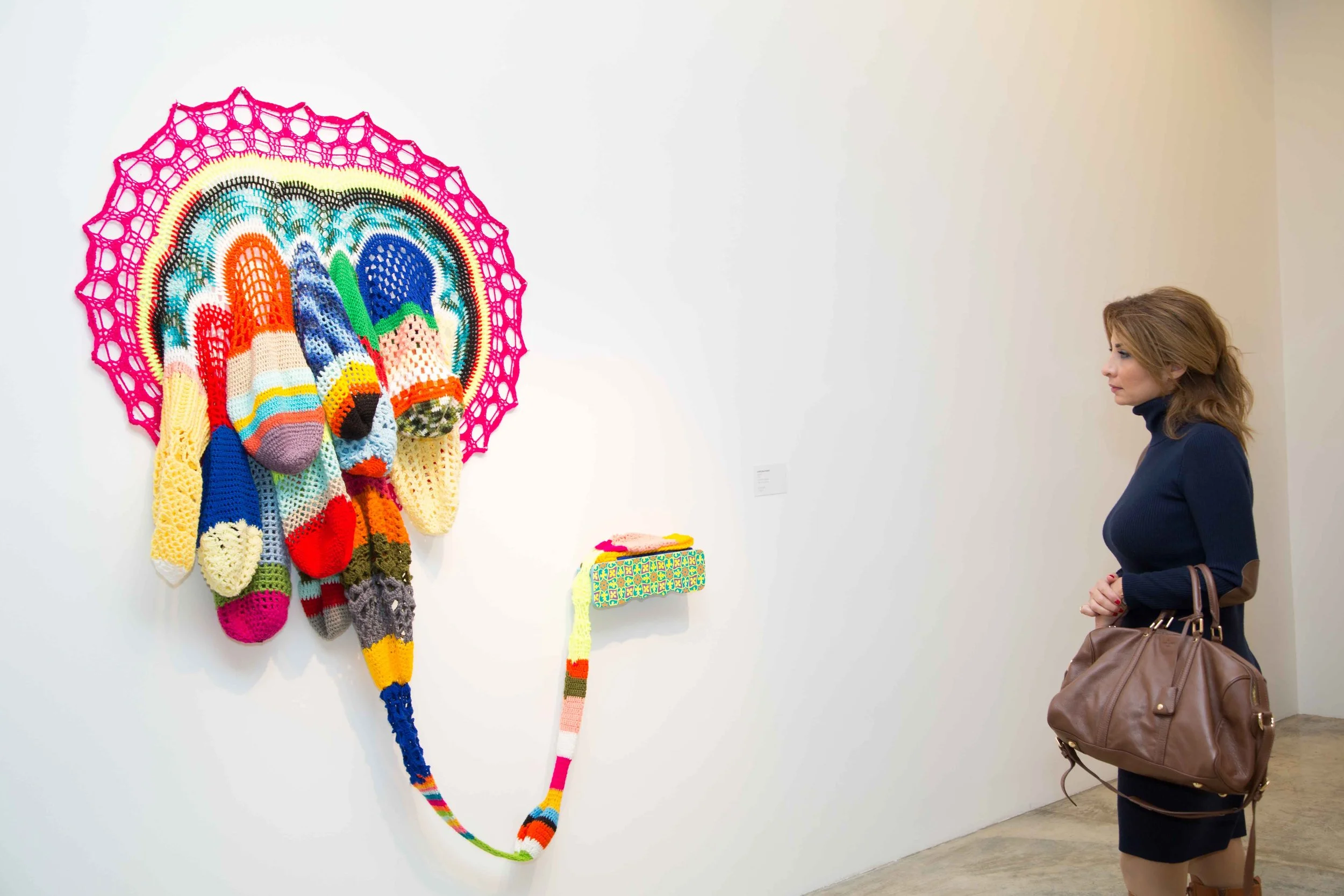 A woman is viewing a colorful, knitted art piece on a white gallery wall. The art includes a large, rainbow-colored circular frame with various knitted items hanging from it, resembling a whimsical face or mask with knitted socks and a long knitted t