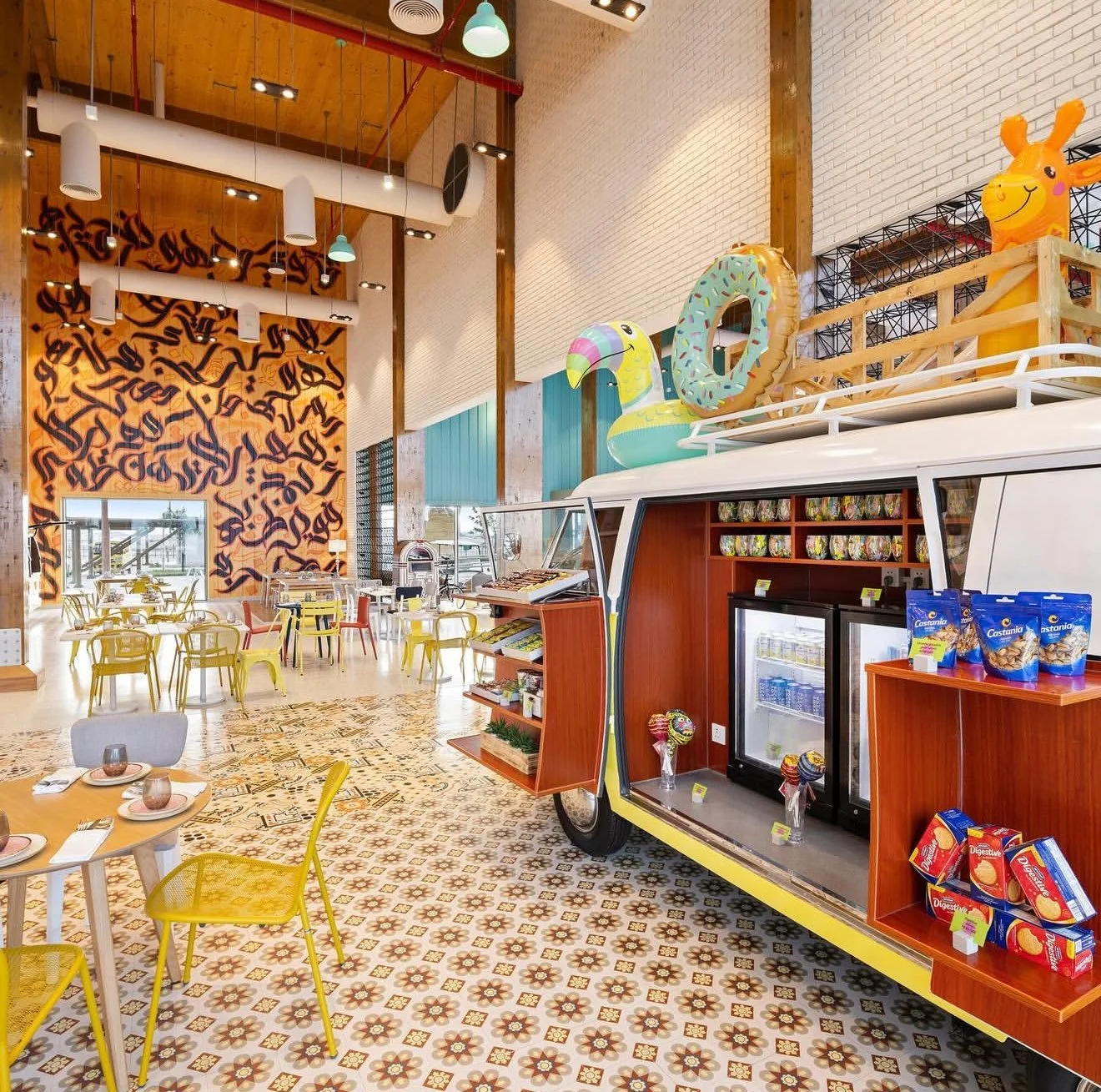 Interior of a brightly colored cafe or ice cream shop with a vintage van serving as a display and counter. The van is decorated with inflatable animal and candy-shaped toys on top. The floor features patterned tiles, and there are tables with yellow 