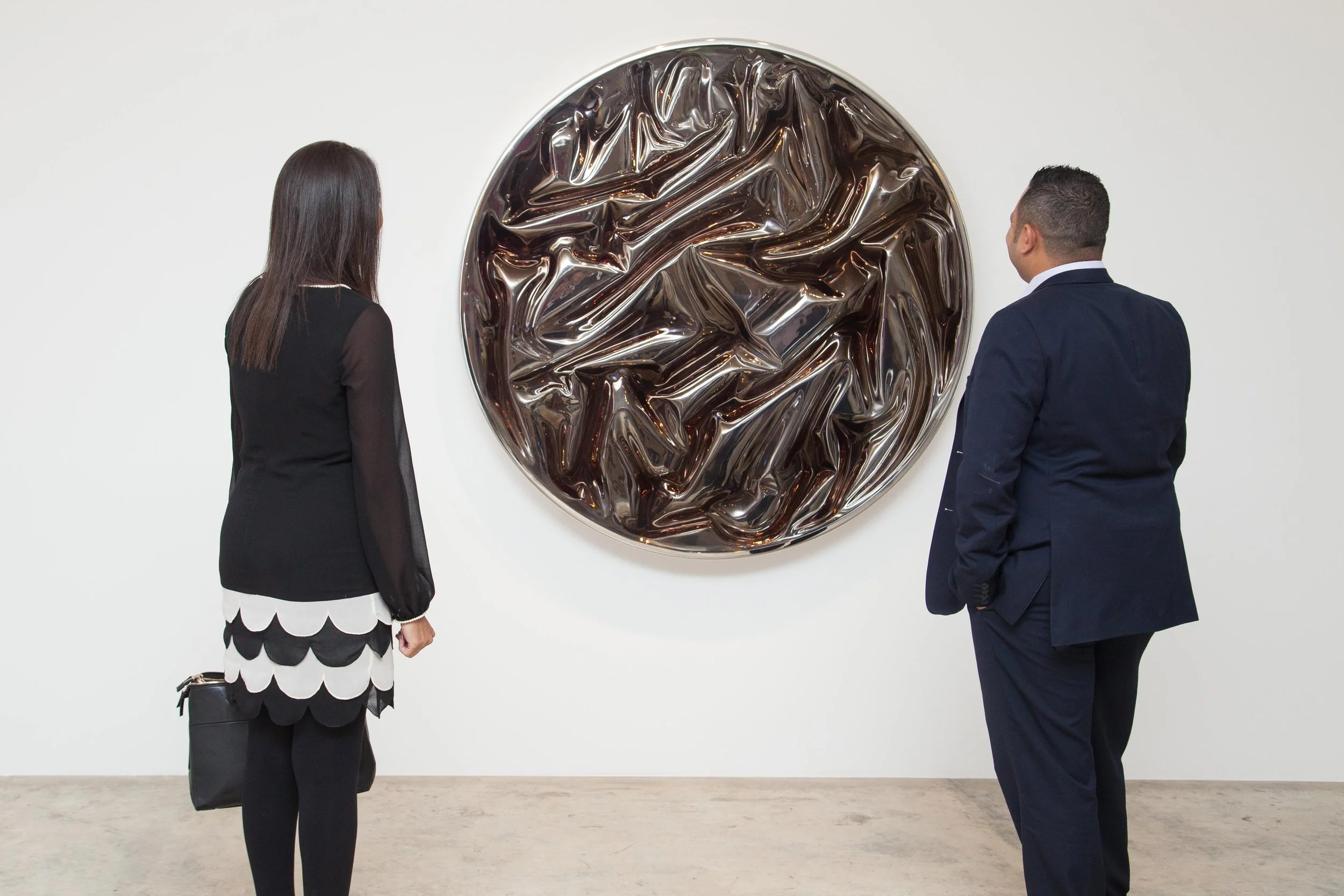 Two people, one woman and one man, stand in front of an abstract round artwork with a crinkled, metallic dark surface, hanging on a white gallery wall.