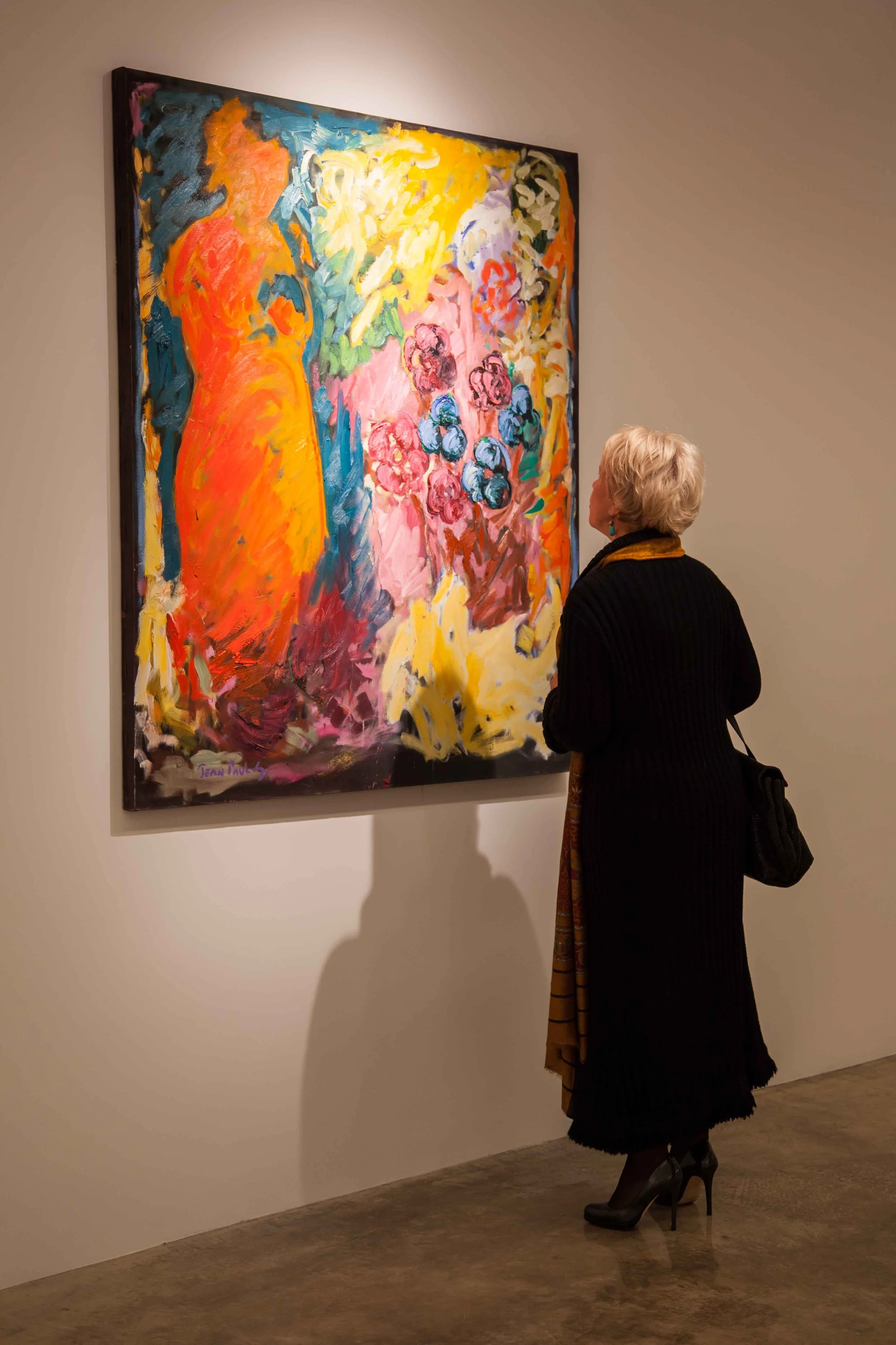 A woman with white hair dressed in black with high heels and a bag looks at a colorful abstract painting of flowers in a gallery.
