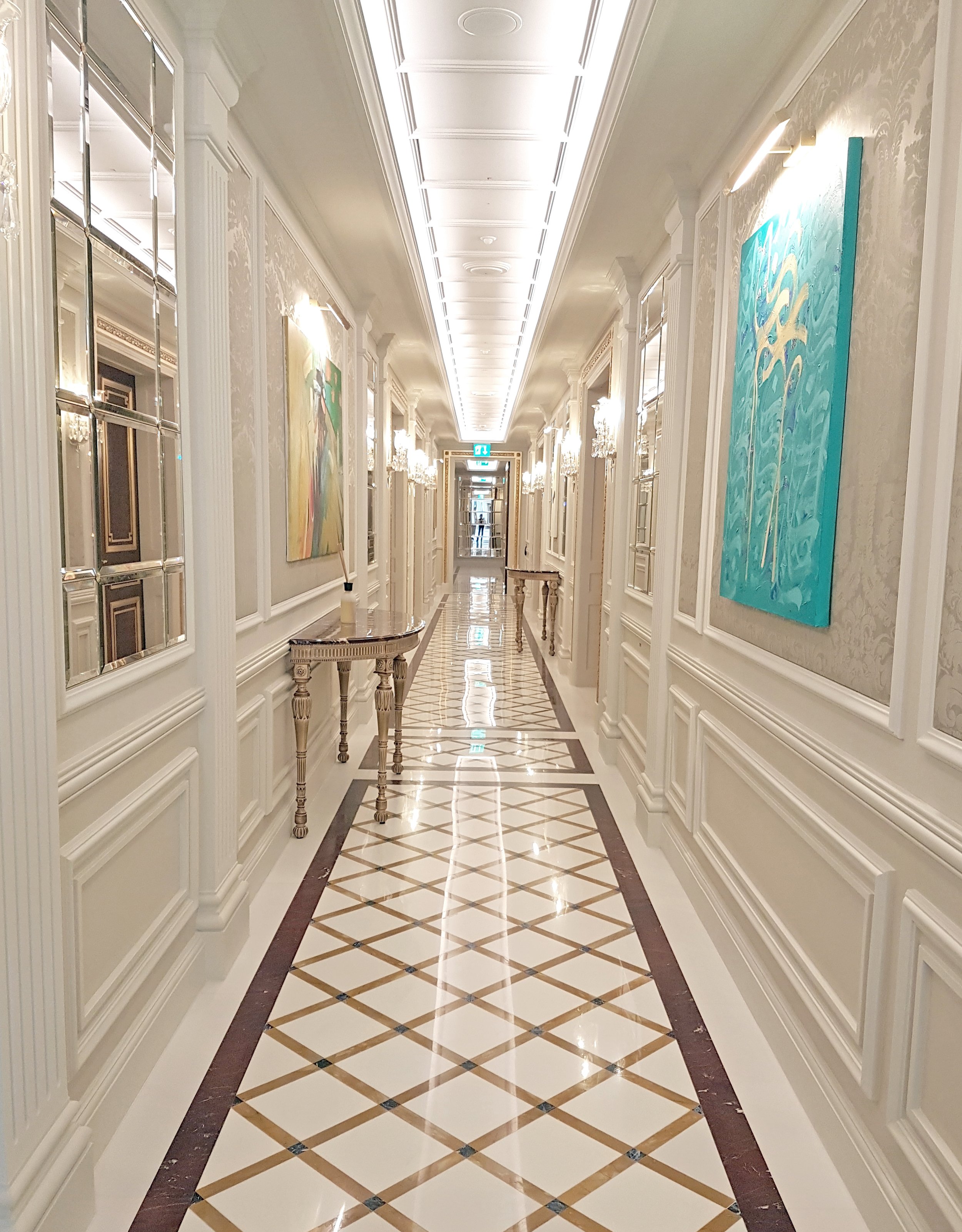 Elegant hotel corridor with white paneled walls, chandeliers, and colorful artwork, leading to a glass door at the end.