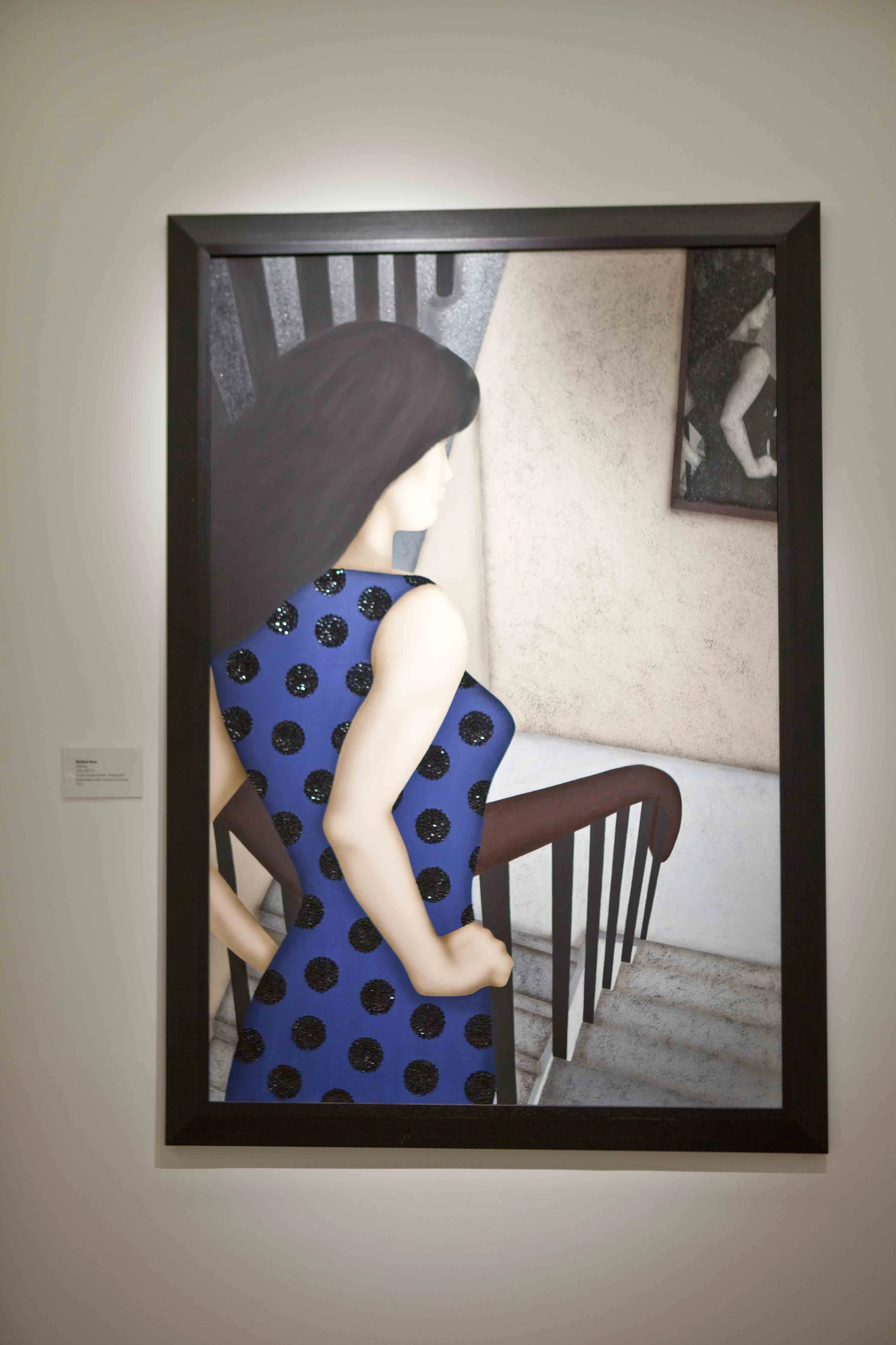 Painting of a woman with dark hair wearing a blue dress with black polka dots, seen from the side, standing on a staircase with a framed picture on the wall in the background.