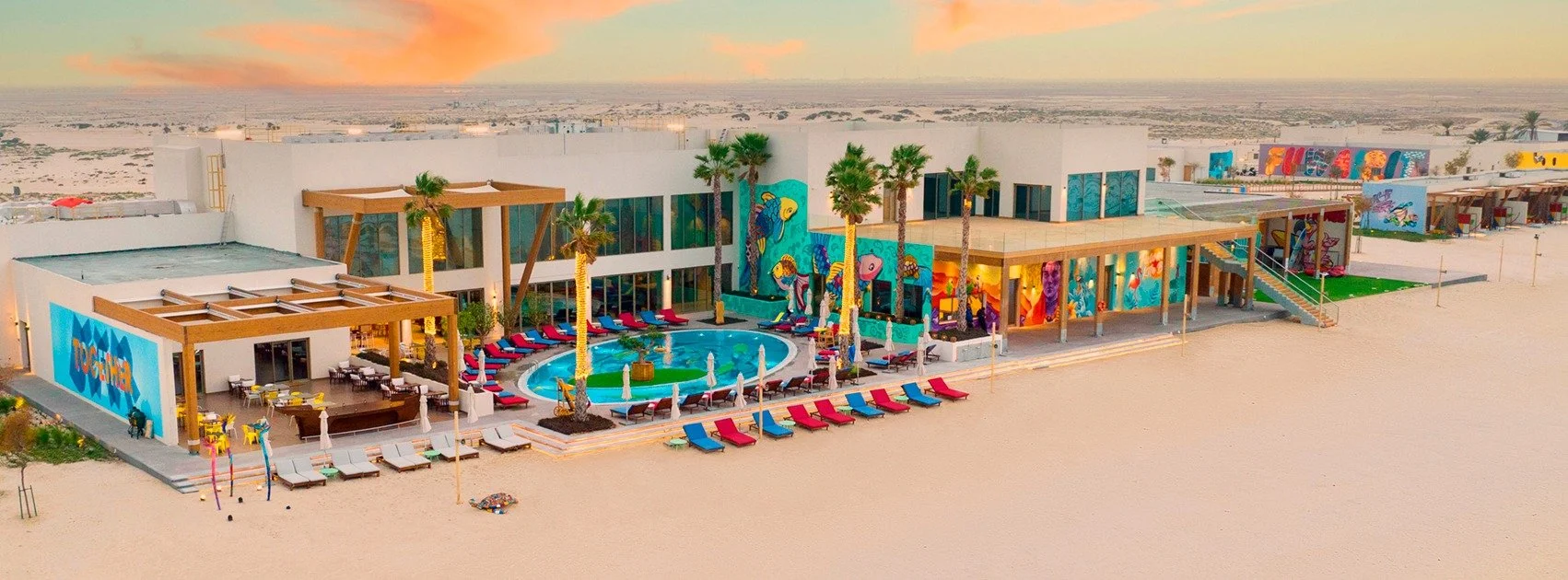Aerial view of a modern hotel with colorful murals, a swimming pool with lounge chairs, and palm trees, located in a desert area during sunset.