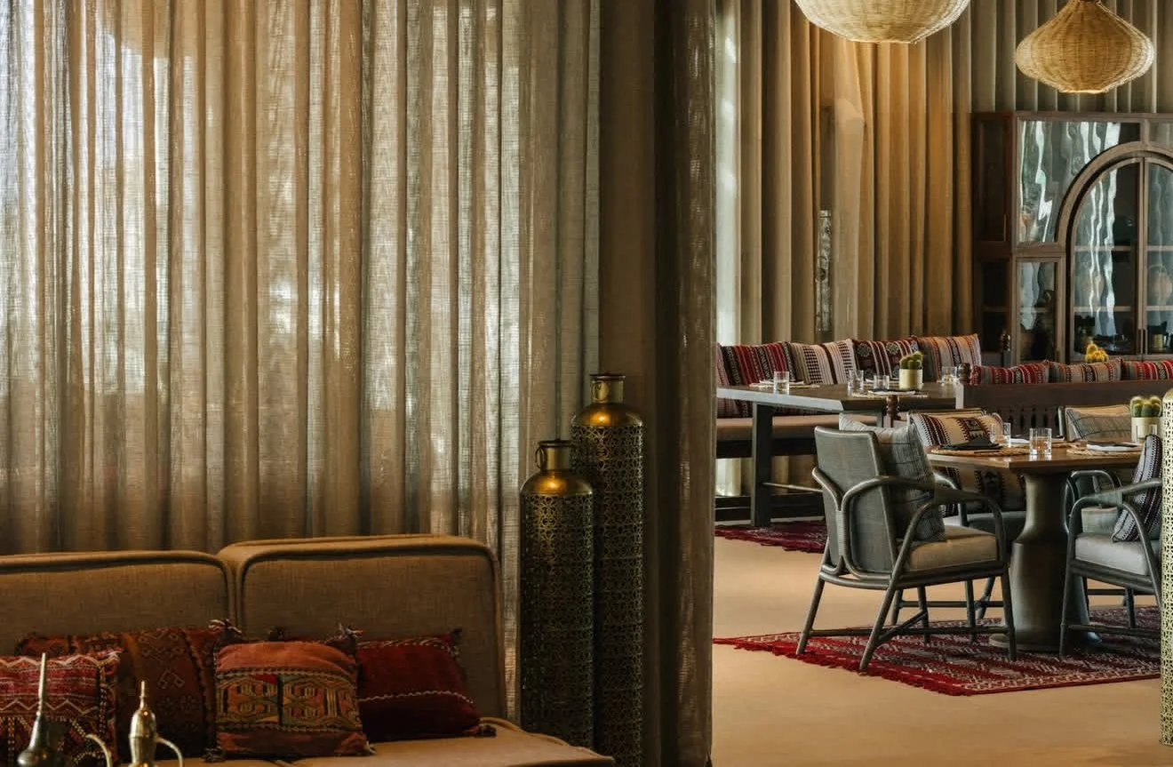 A cozy restaurant interior with curtains, a sofa with decorative pillows, a table set for dining, and woven pendant lights.