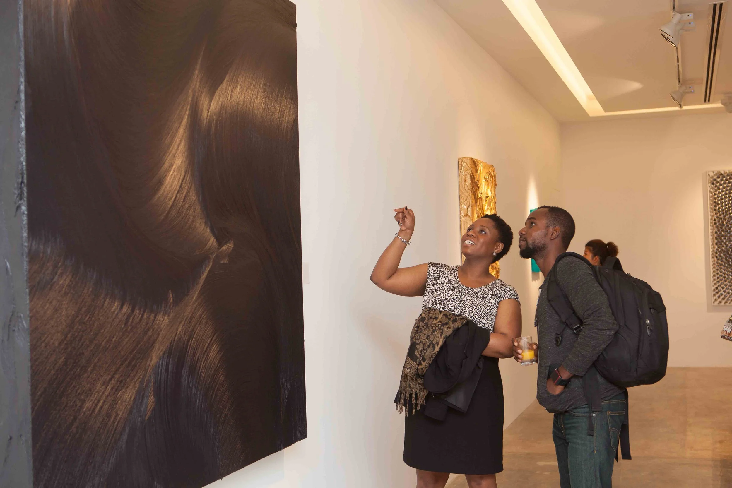 A woman and a man are looking at artwork in an art gallery. The woman is gesturing towards the painting with her right hand while holding a jacket and purse. The man is observing the artwork with a backpack on his back, holding a drink in his right h