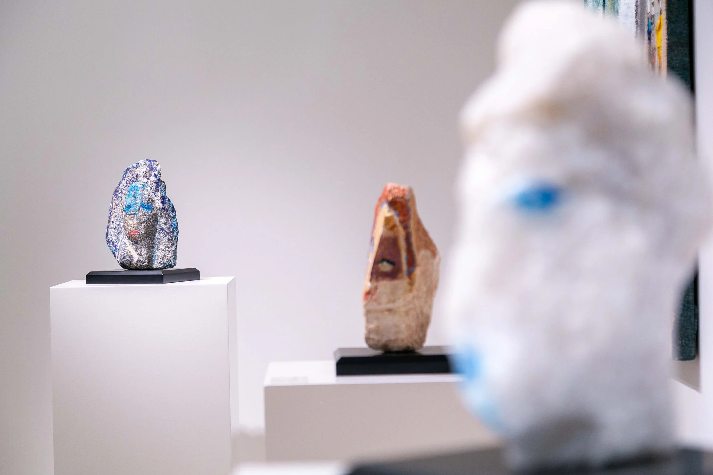 Three sculptures on display in an art gallery. The sculpture on the left features a face with blue hair, closed eyes, and a neutral expression. The middle sculpture resembles a face with closed eyes and a brownish-orange tone. The sculpture on the ri