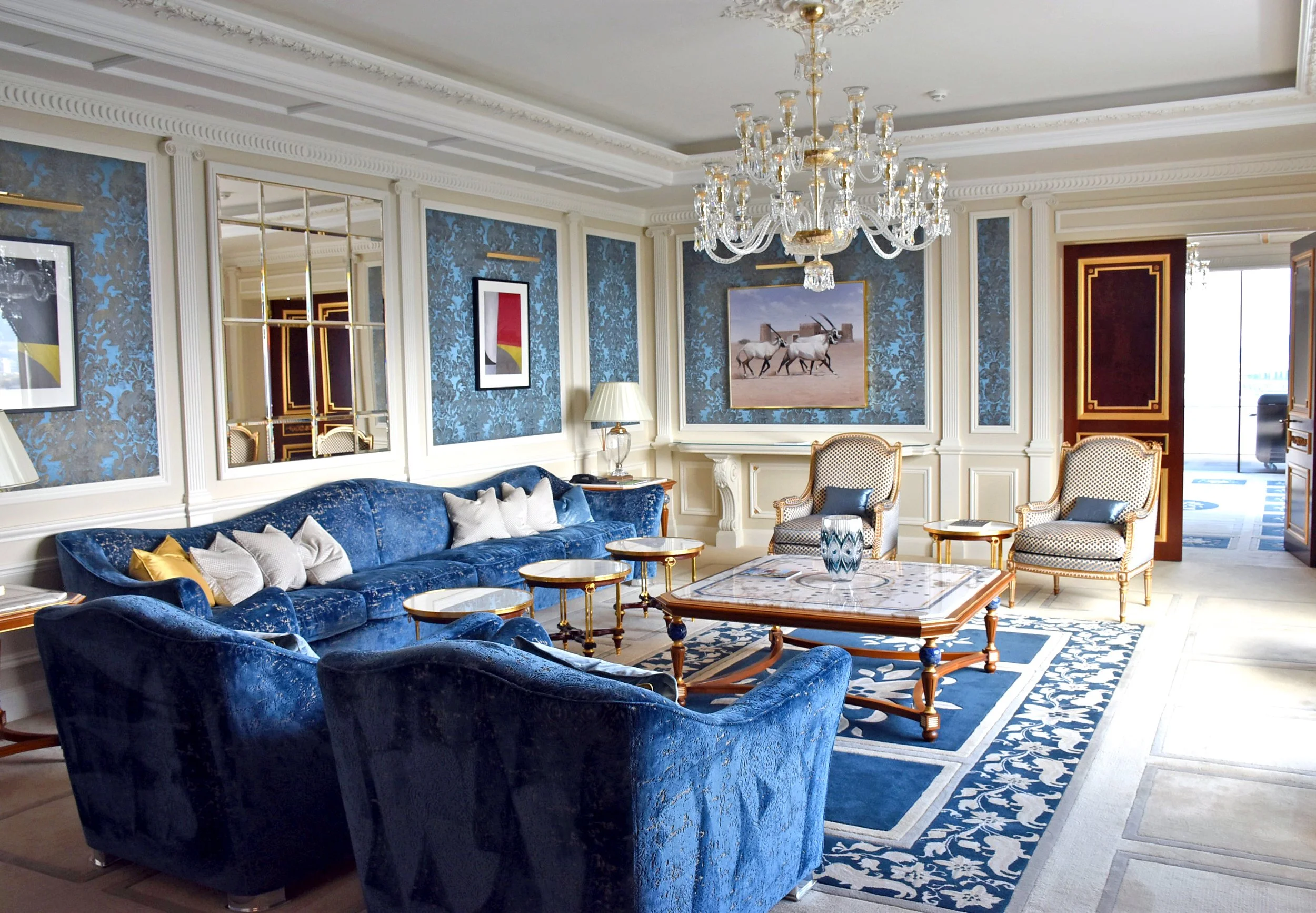 Luxurious living room with deep blue velvet sofas, patterned armchairs, a marble-topped coffee table, gold accents, framed artwork, a chandelier, and elegant wall molds and wallpaper.