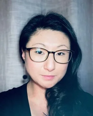 A woman with long black hair and glasses looking slightly to the side against a gray background.